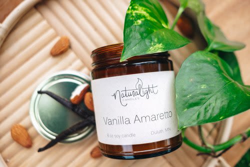 Brown glass Vanilla Amaretto candle jar with white label on a tray beside almonds, vanilla beans, and a green vine.