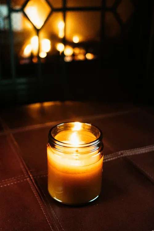 Lit amber candle in a small glass jar on a dark surface with warm light in the background
