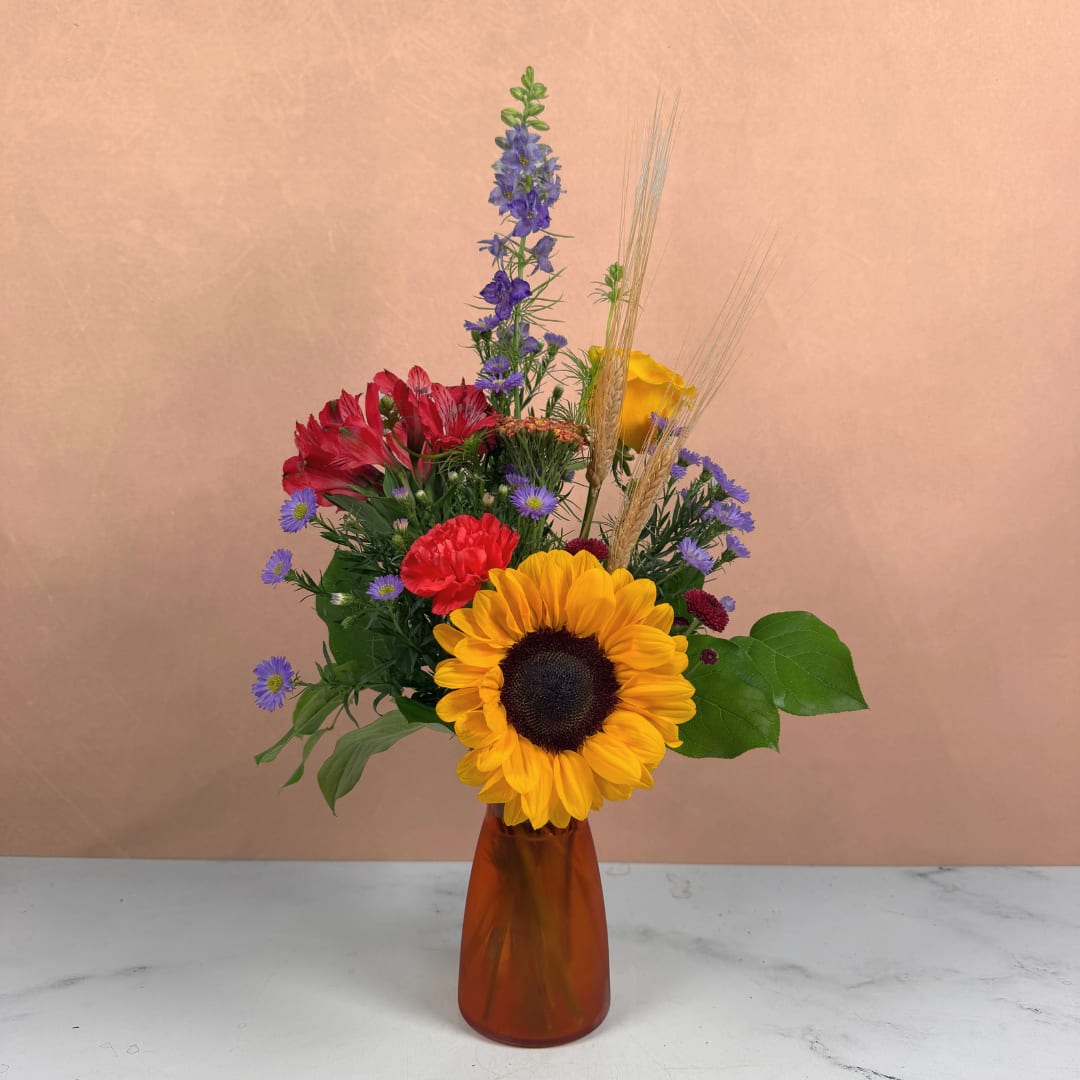 Mixed bouquet with a sunflower, red blooms, and purple flowers in an amber vase