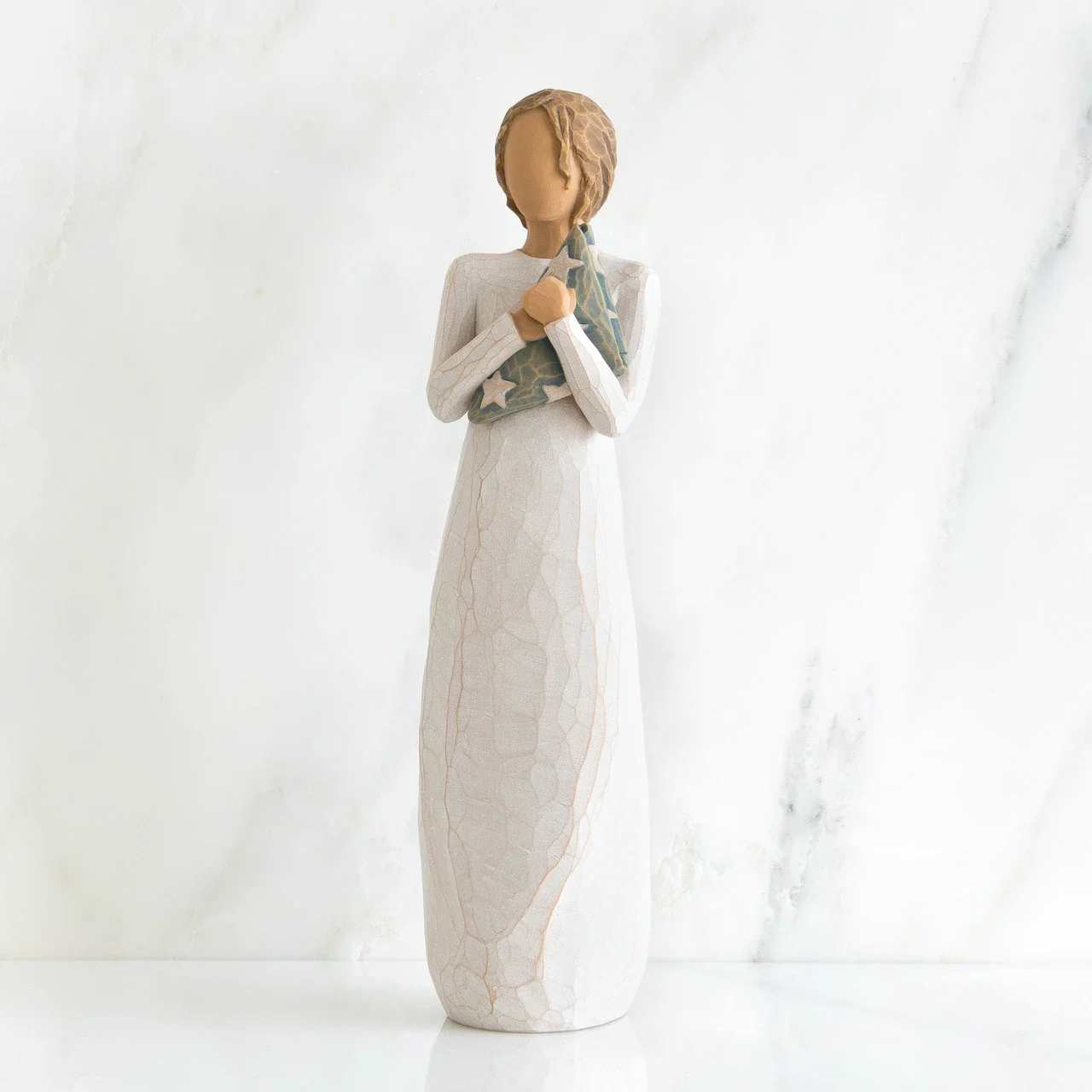 Willow Tree Hero - A quiet keepsake with a soft, hand-carved feel, this Willow Tree Hero figurine is a tasteful sympathy or memorial gift and a gentle choice for expressing care after a loss. The tall, faceless figure in a long white dress holds small blue flower bundles (the only vivid color), creating a calm, contemplative focal point that reads as home décor and remembrance rather than a floral arrangement.  Hand-finished with muted beige skin tones and textured white drapery, the piece feels like a handcrafted bedside or mantel accent-comforting to touch and soothing to view. The muted blue-green star-patterned shawl detail in some versions adds subtle character. Perfect for placing on a dresser, memorial shelf, or beside a photo, this keepsake offers a simple way to let the person you're thinking of know they're not alone.