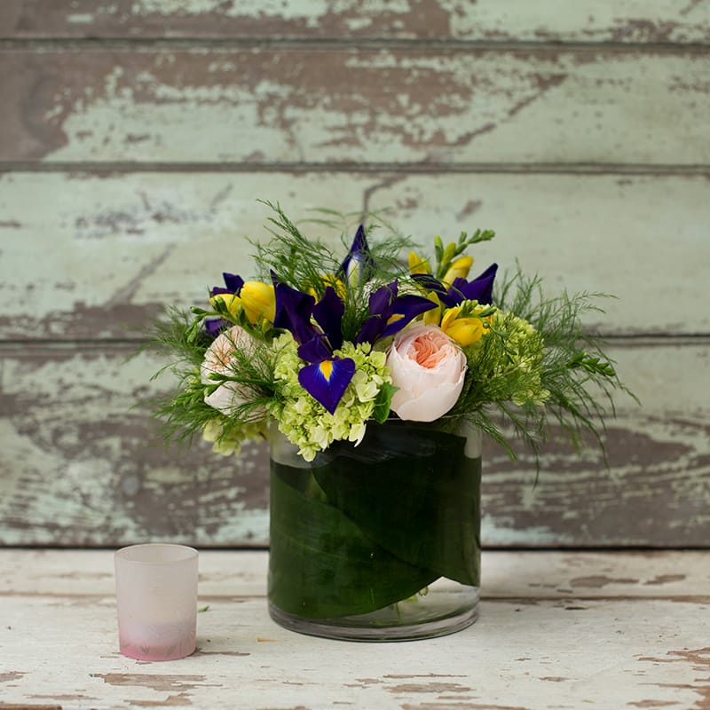 Bouquet of purple irises, yellow blooms, and pale pink roses in a glass vase