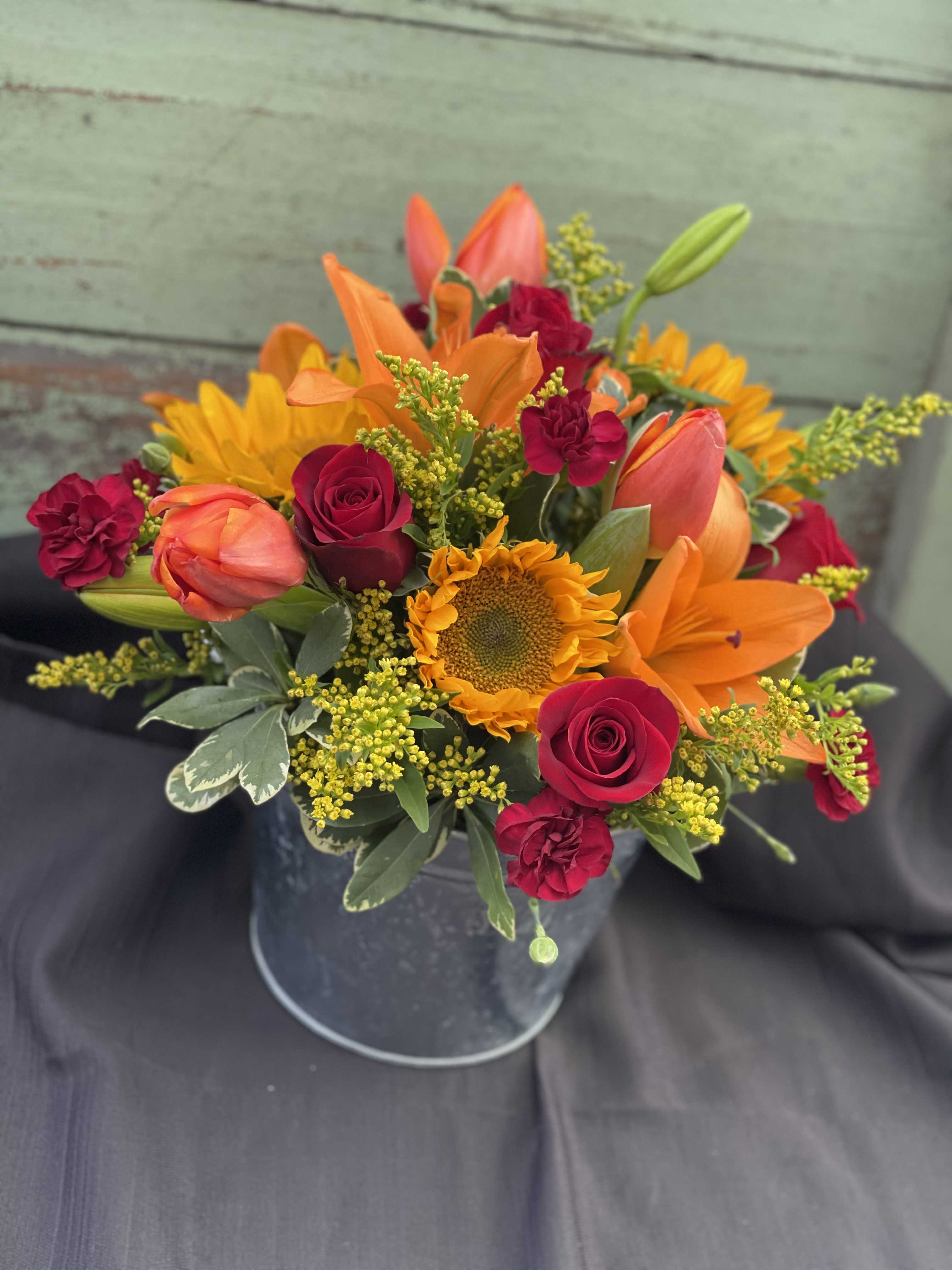 Bouquet of orange lilies, red roses, and a sunflower in a metal bucket