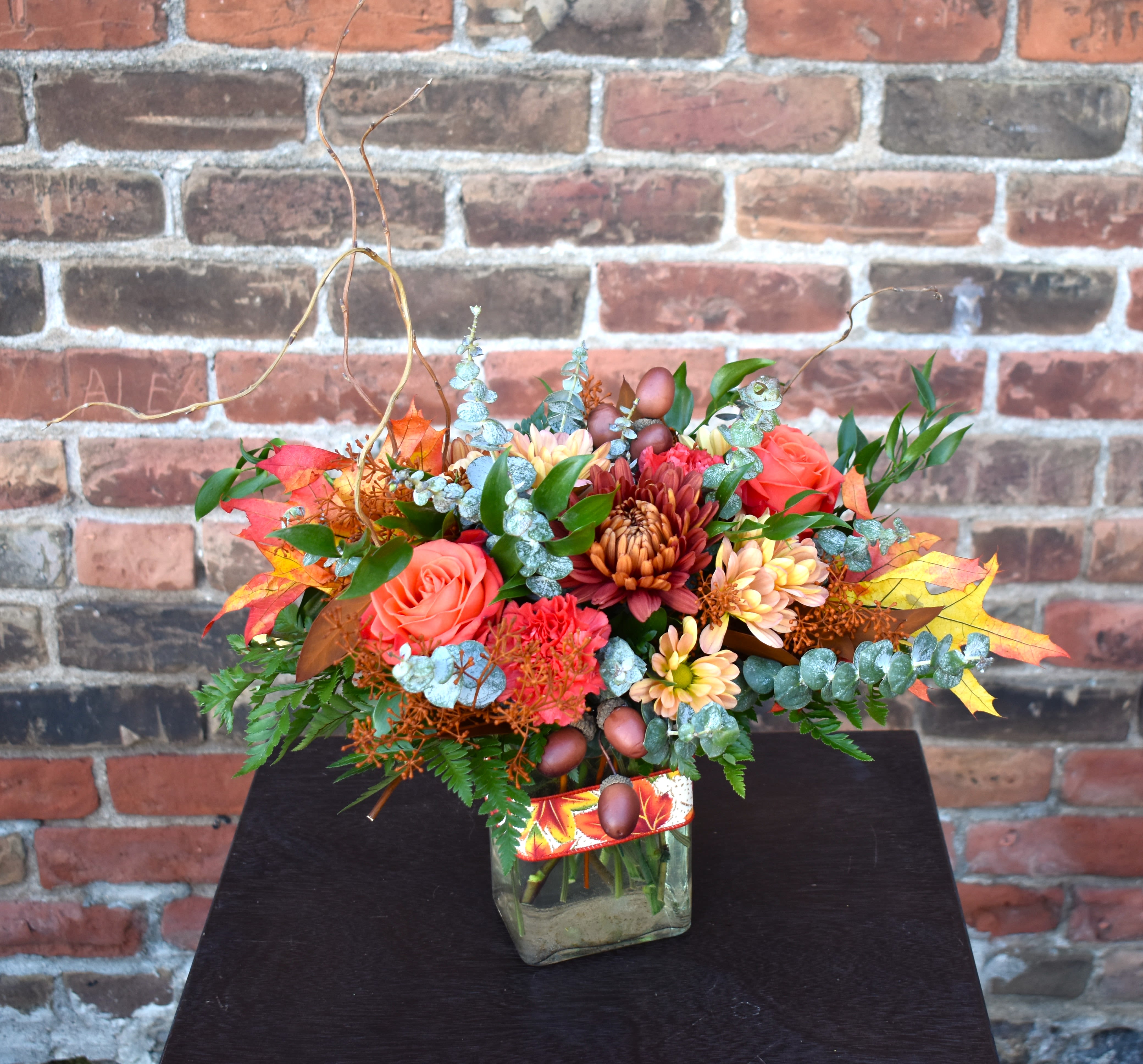 Acorn - Brilliant fall hues fill this bouquet! Bright orange roses, fall mums, orange carnations, curly willow, and a mix of eucalyptus are designed in a clear vase and accented with fall leaves and acorns...the perfect way to celebrate everything this season has to offer! 