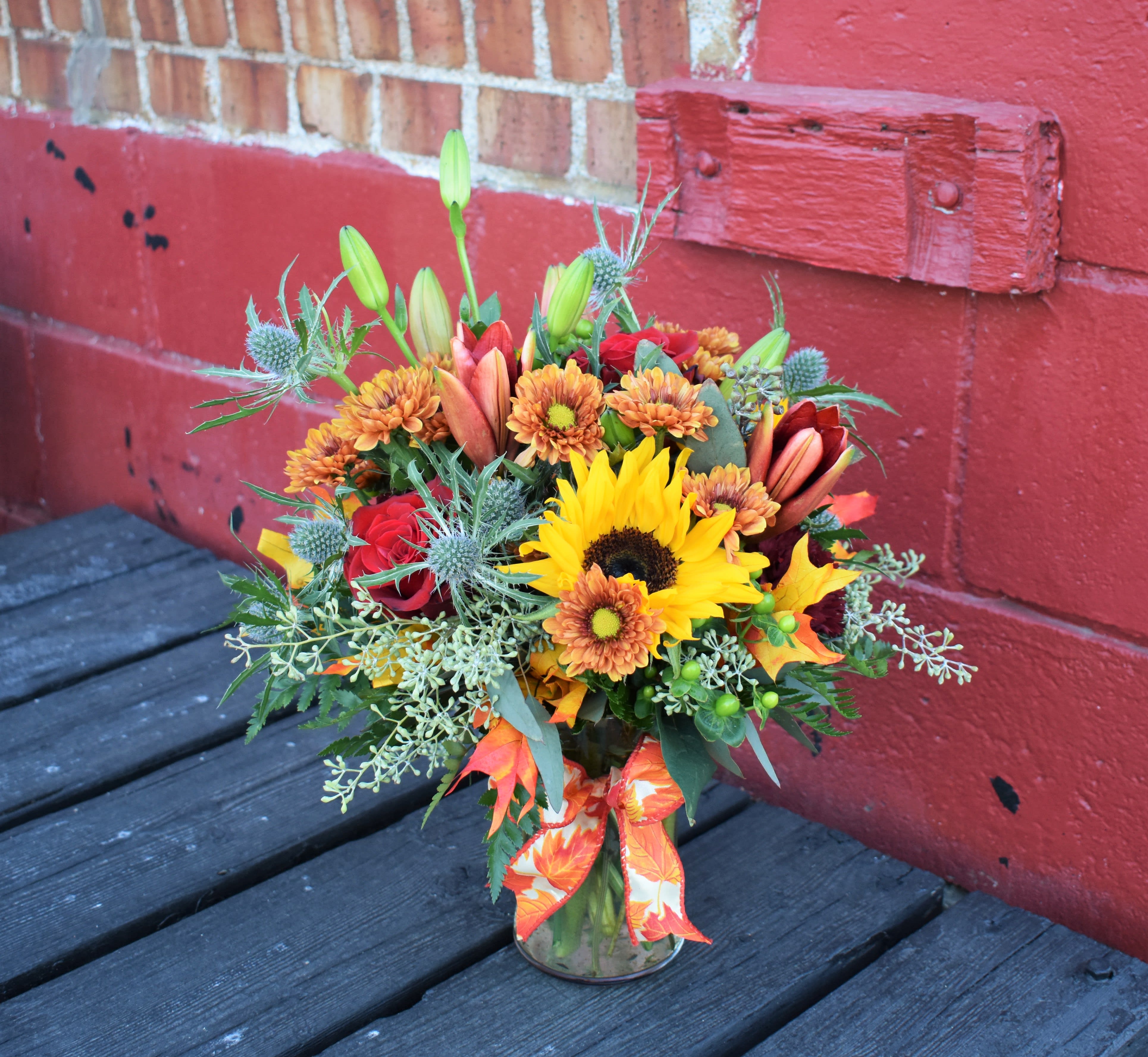 All For Fall - This seasonal arrangement of burgundy lilies, red roses, butterscotch mums, bright sunflowers, blue thistle and eucalyptus is designed in a glass vase and accented by fall leaves and a cute fall bow...the perfect way to celebrate any fall occasion! 