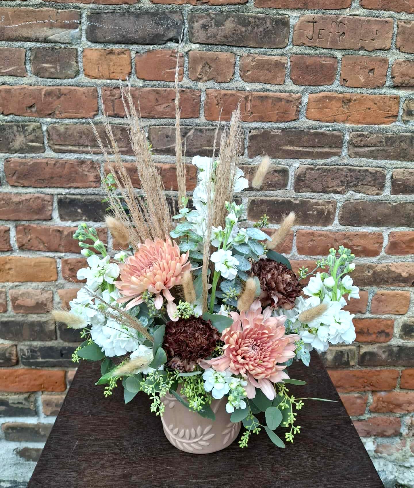 Crème Café - A toasty-warm seasonal design created in neutral toned, leaf-accented pottery, featuring white stock, peach mums, copper carnations, eucalyptus, and accented with rustic grasses...the perfect seasonal design to celebrate any occasion!  Approx 1.5 ft X 1 ft
