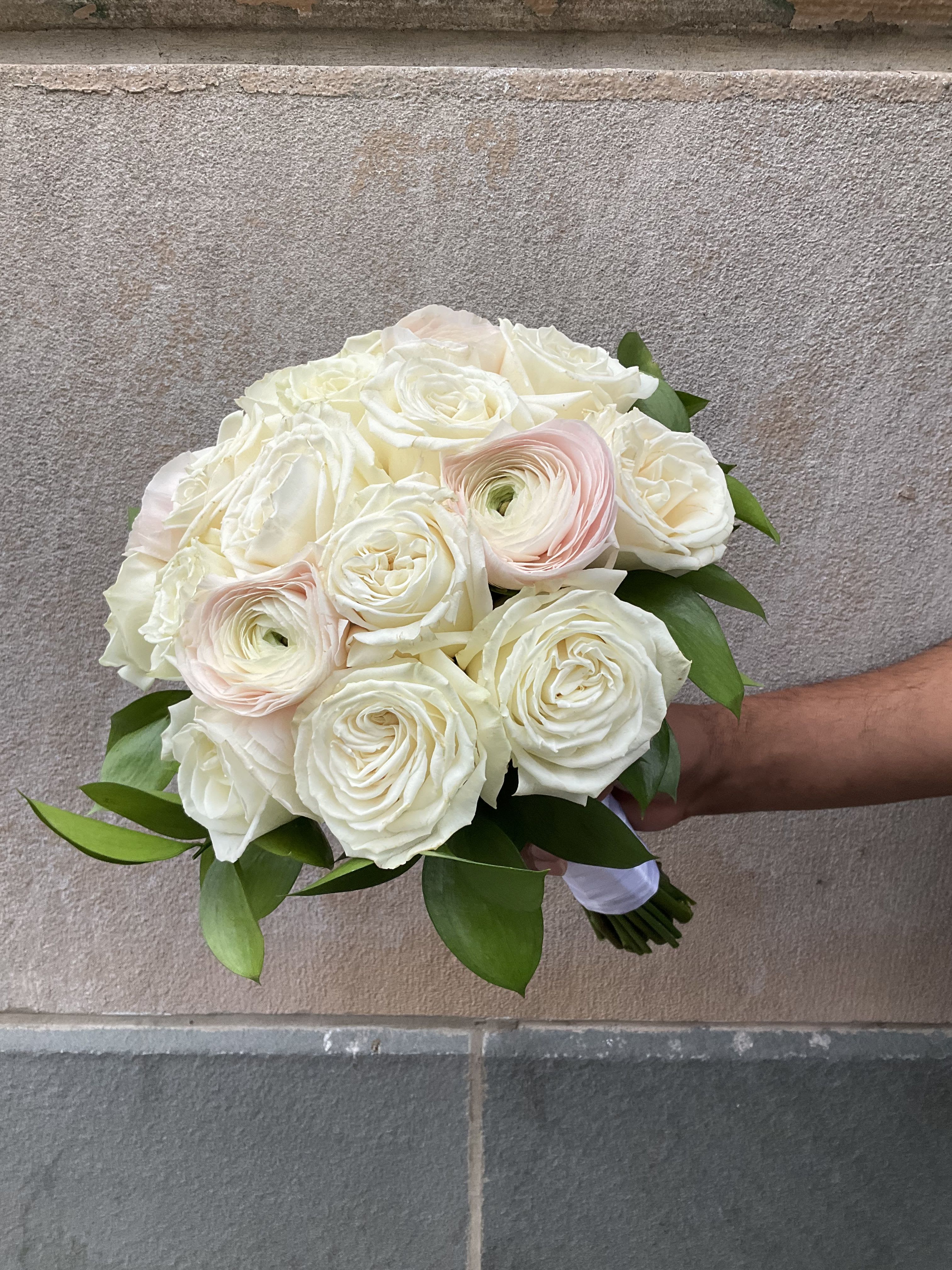 Pure Grace Hand-Tie - A beautifully hand-tied bouquet designed in a tight, compact style, where each bloom is carefully arranged with the heads close together for a clean and elegant look. Featuring 14 roses and 4 ranunculus, this bouquet highlights soft texture and fullness while maintaining a refined, structured shape. Finished with a ribbon-wrapped handle, it is perfect for carrying, gifting, or celebrating any special moment with style.  This bouquet is available in two distinct styles: one surrounded by soft baby’s breath for a classic, airy look, or one styled with fresh greenery for a more natural, modern feel. You can choose the option that best fits your preference in the notes when ordering the flowers.  Please note that flower varieties may vary slightly based on daily availability. We always do our best to maintain the same look, color palette, and overall design using the freshest flowers available.