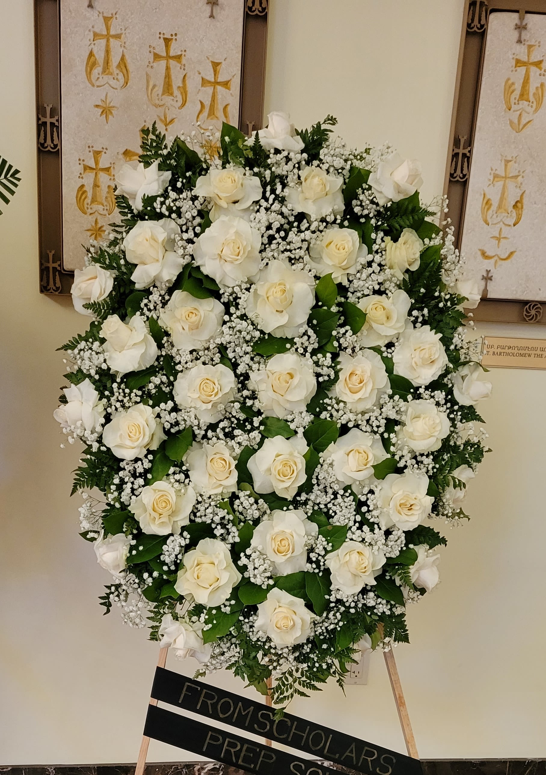 Oval standing spray of white roses and baby's breath on an easel with a black dedication sign.