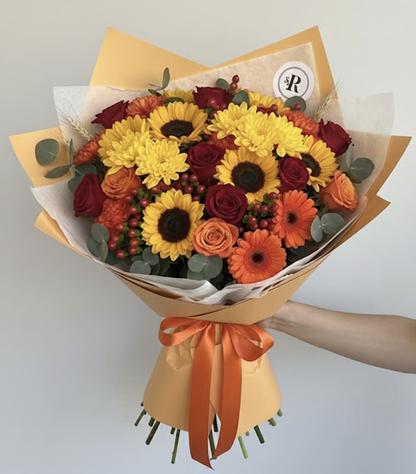 Hand-tied bouquet of yellow sunflowers, red roses, and orange blooms in orange wrap with ribbon