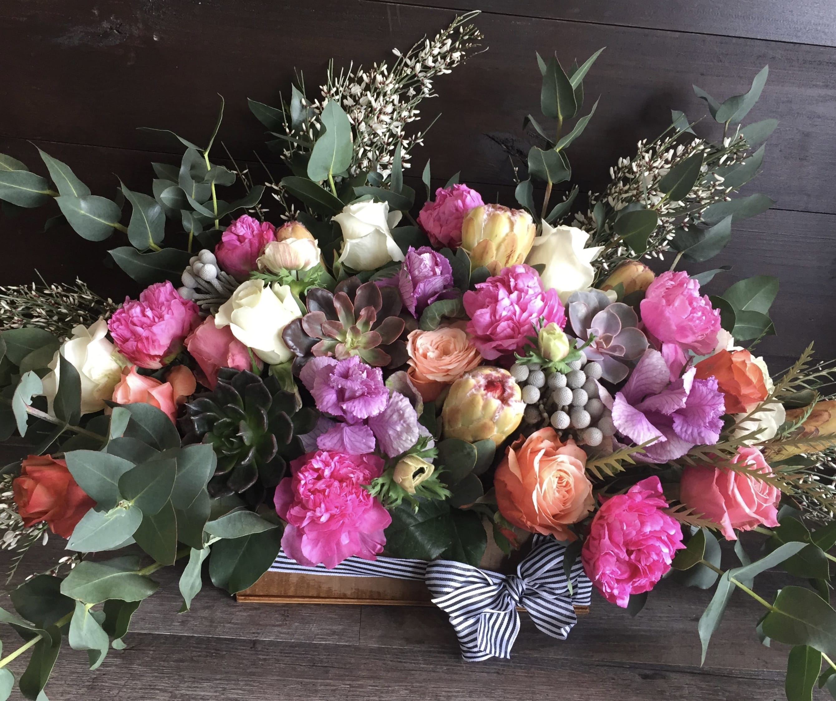 Low rectangular arrangement of pink peonies, mixed roses, succulents and protea in a wooden box with a striped bow