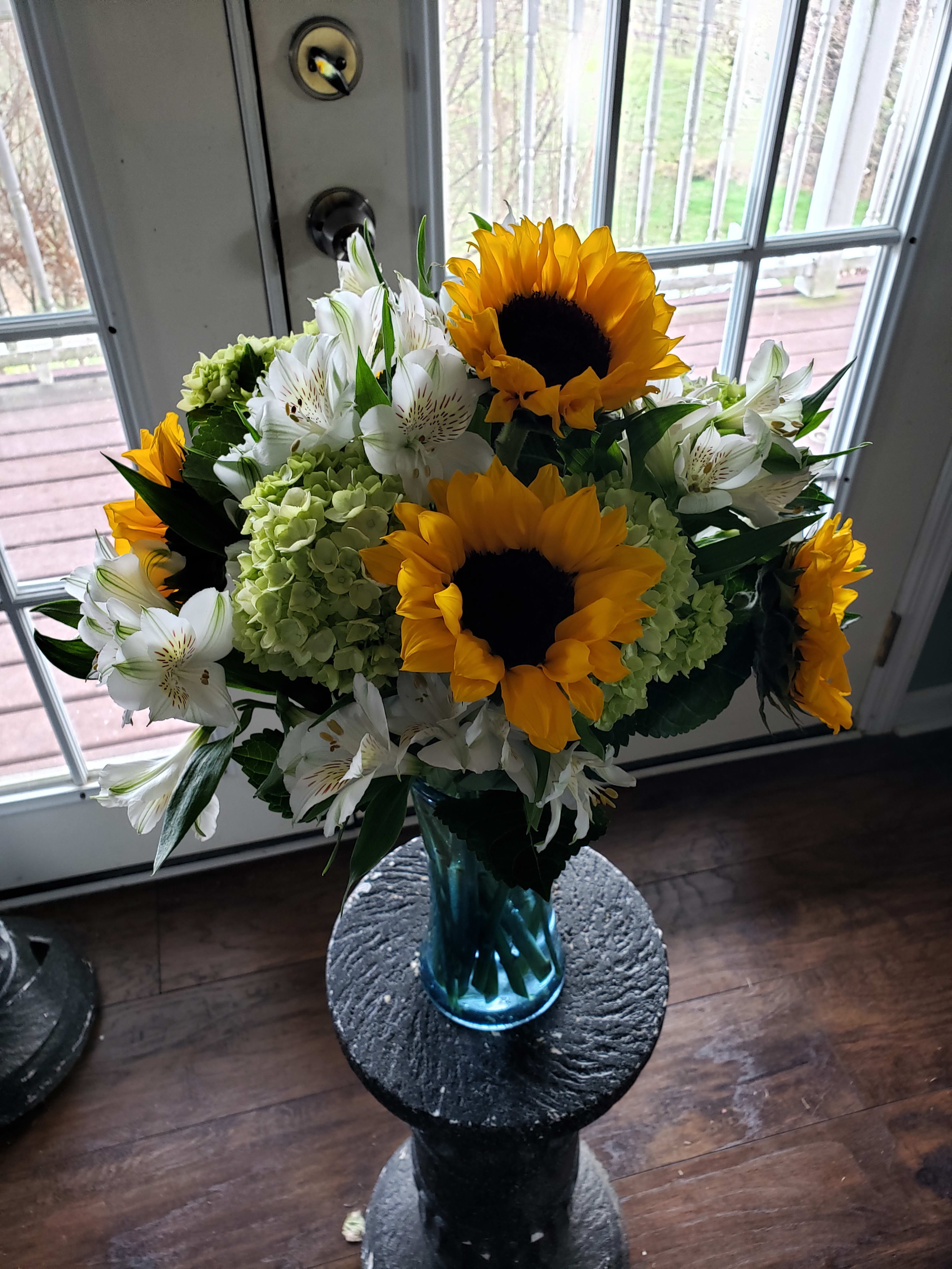 Sunny Day Sunflower Arrangement - Brighten someone's day with this beautiful summer inspired floral arrangement. Fresh farm grown sunflowers arranged in a blue glass vase accented with white flowers and seasonal greenery.