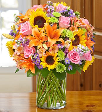 Mixed bouquet of sunflowers, pink roses, orange lilies, and purple flowers in a glass vase