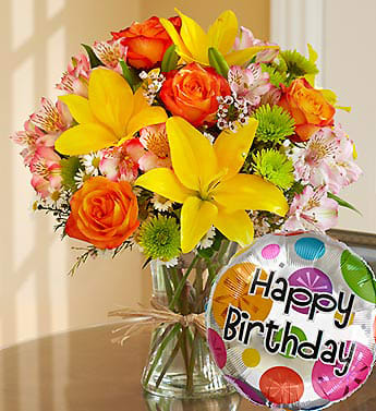 Bouquet of yellow lilies, orange roses, and pink flowers in a glass vase with a birthday balloon.