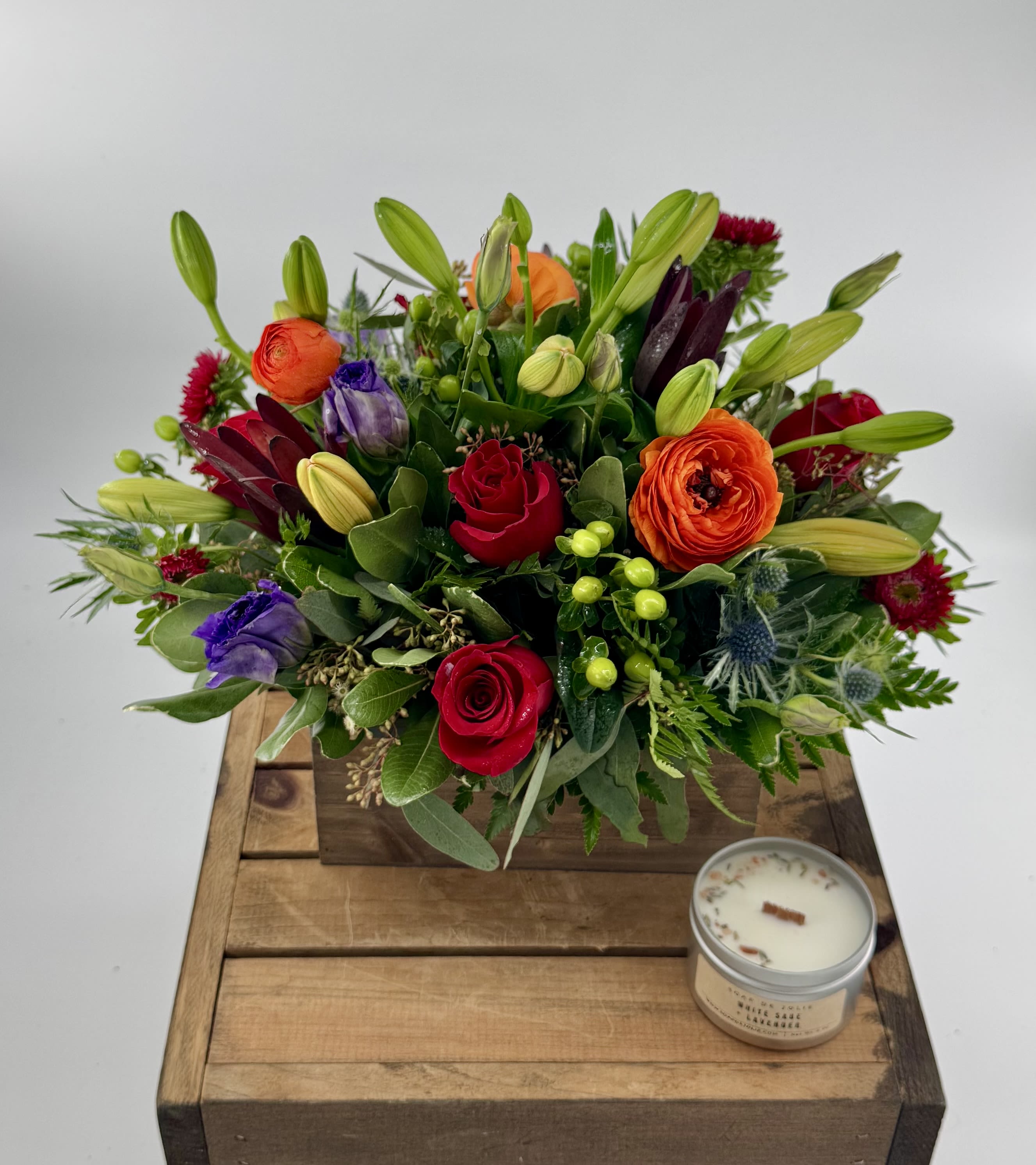Mixed bouquet with roses, lilies, and purple blooms in a wooden box beside a candle