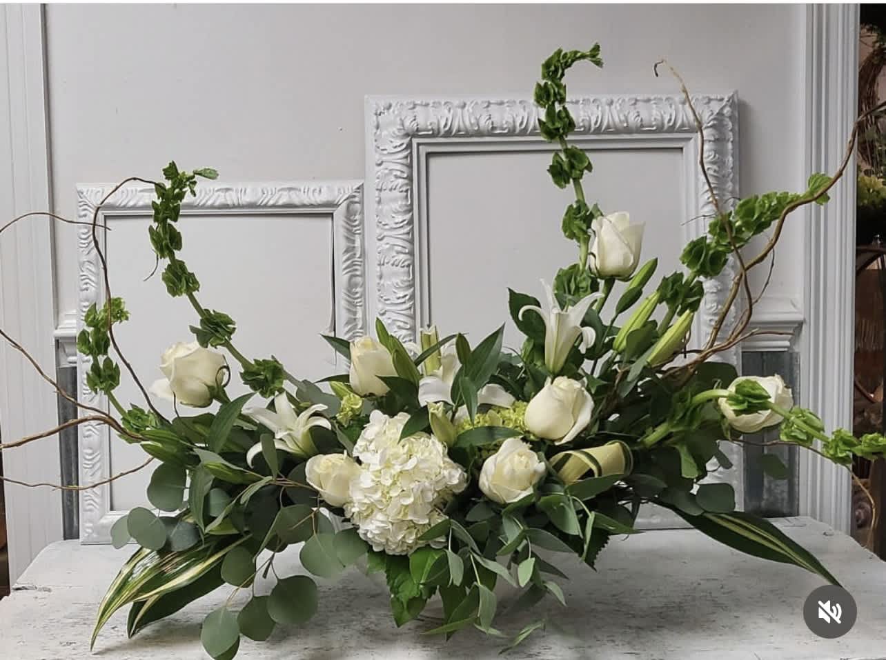 Long low arrangement of white roses, lilies, and hydrangea with tall green accents