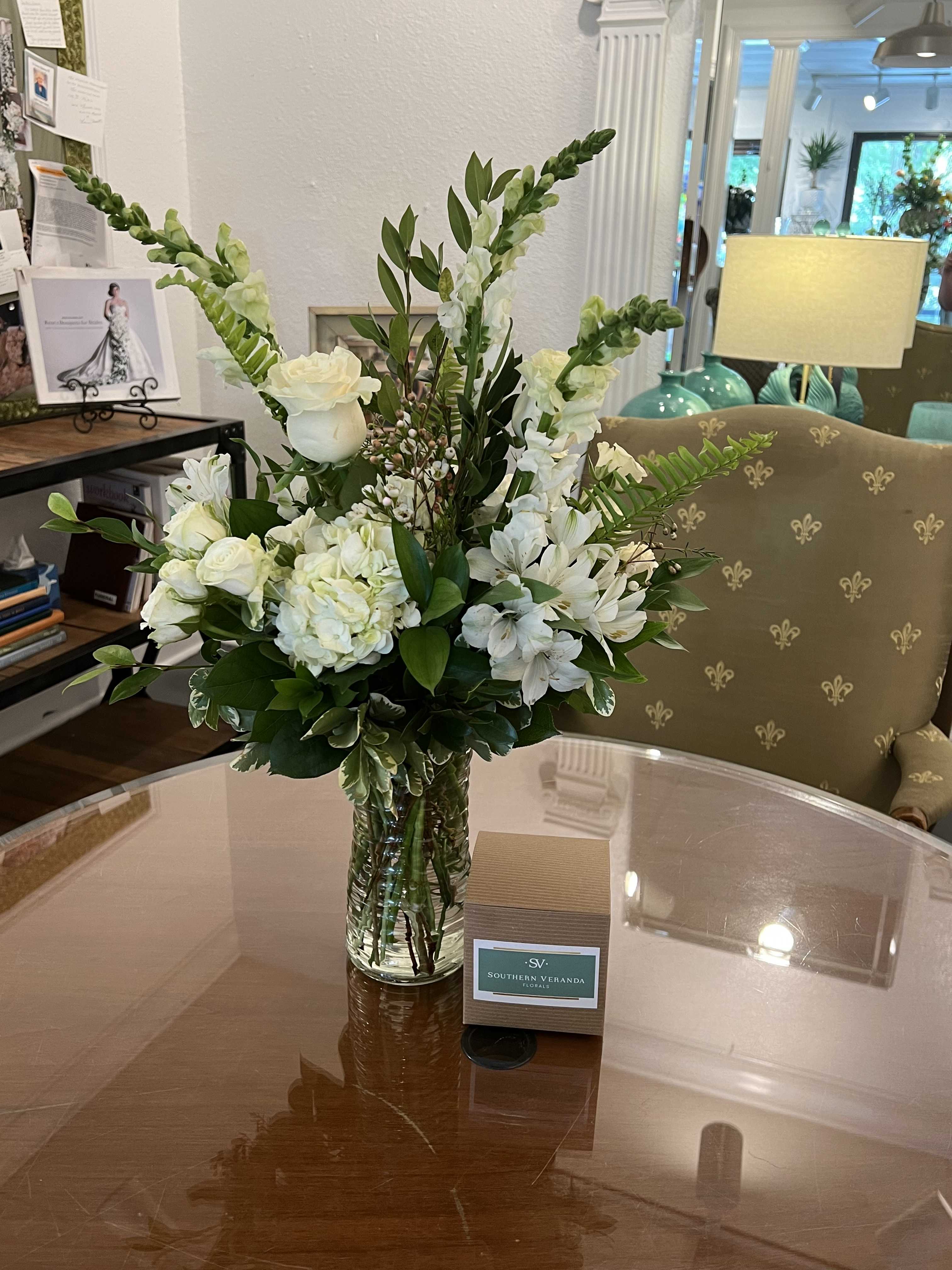 White flower arrangement with roses and snapdragons in a clear glass vase beside a small boxed gift.