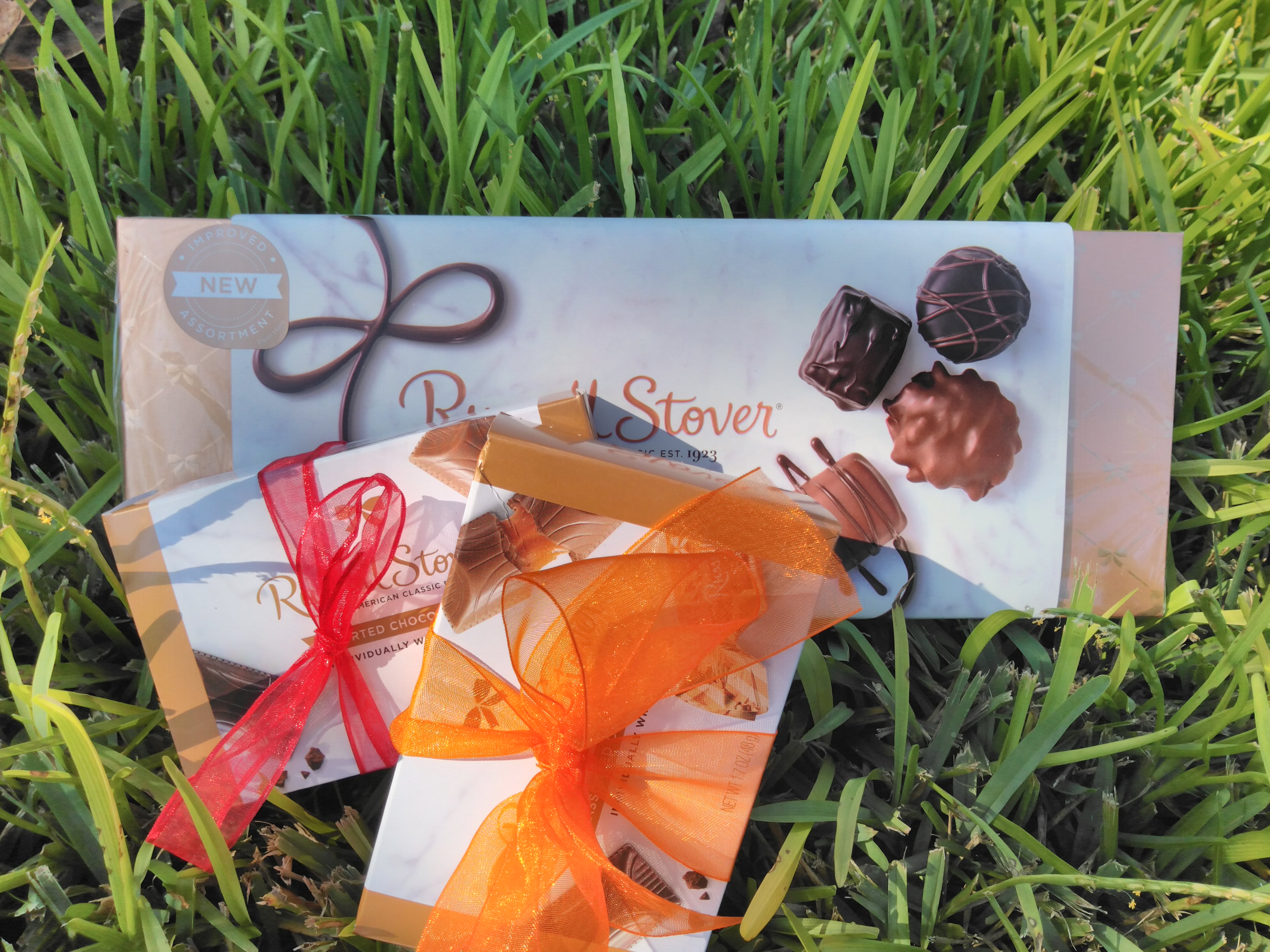 Three boxed chocolate assortments tied with red and orange ribbons on grass.