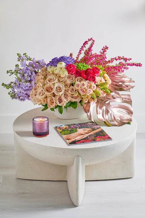 Mixed bouquet of roses and purple flowers in a white vase with a candle and magazine