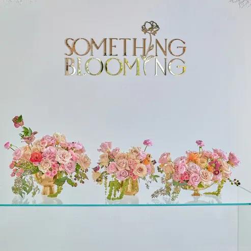 Three pink floral arrangements in gold vases beneath a "Something Blooming" sign