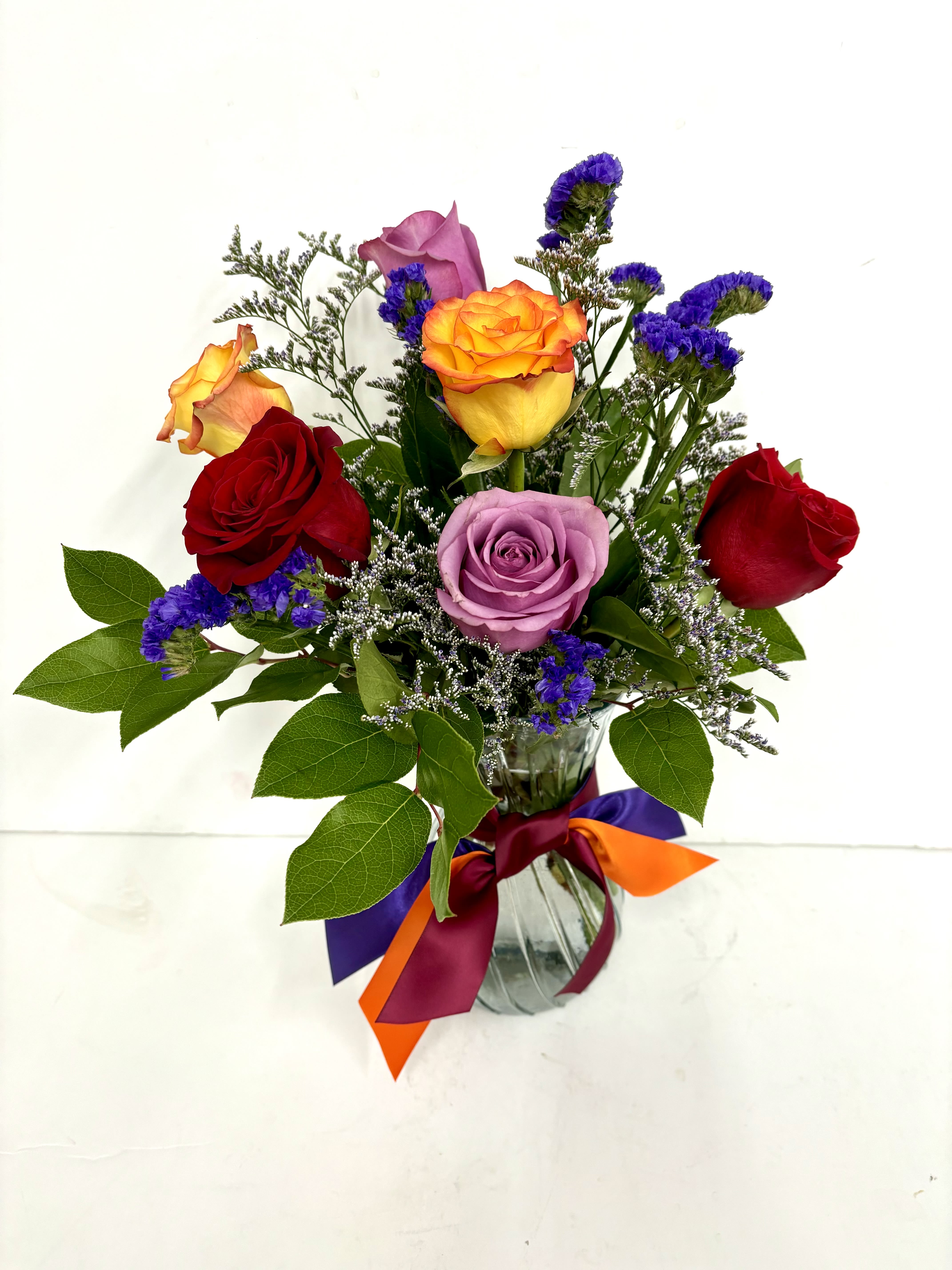 Mixed bouquet of red, yellow, and lavender roses with purple filler flowers in a glass vase with colorful ribbons