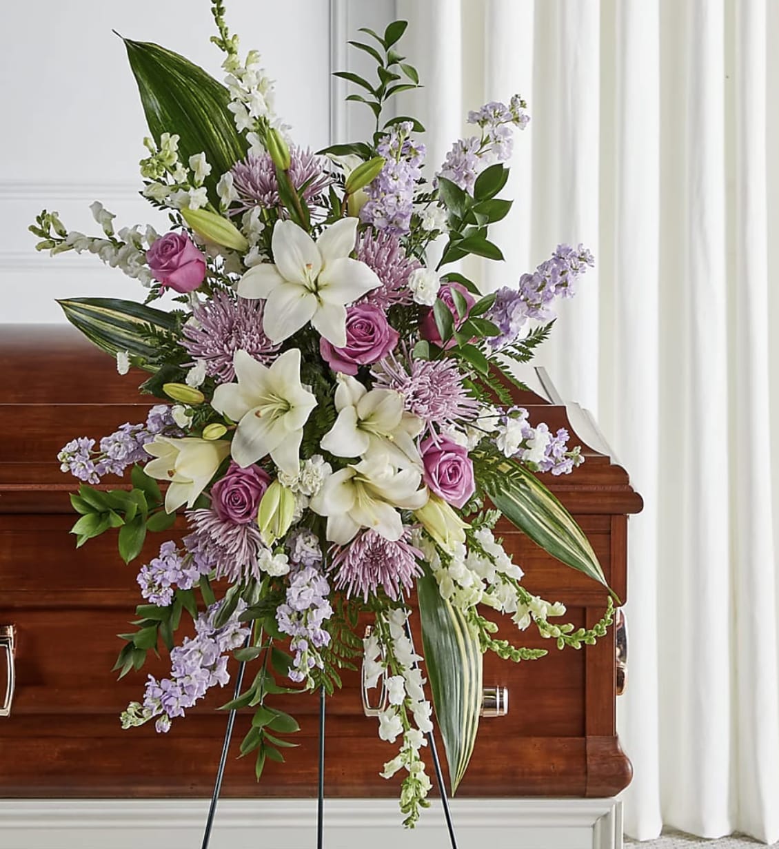 Lavender Grace Standing Spray - Honor a life of beauty, kindness, and love with Lavender Grace Standing Spray. This elegant standing tribute features pristine white lilies, soft lavender roses, delicate spider mums, graceful stock, and premium seasonal blooms, creating a serene expression of comfort and remembrance. Handcrafted by the floral designers at Bloom Luxury Box, each arrangement is thoughtfully designed to celebrate treasured memories while offering heartfelt support to family and friends. Displayed on an easel, this timeless tribute provides a beautiful farewell filled with peace, dignity, and enduring love.