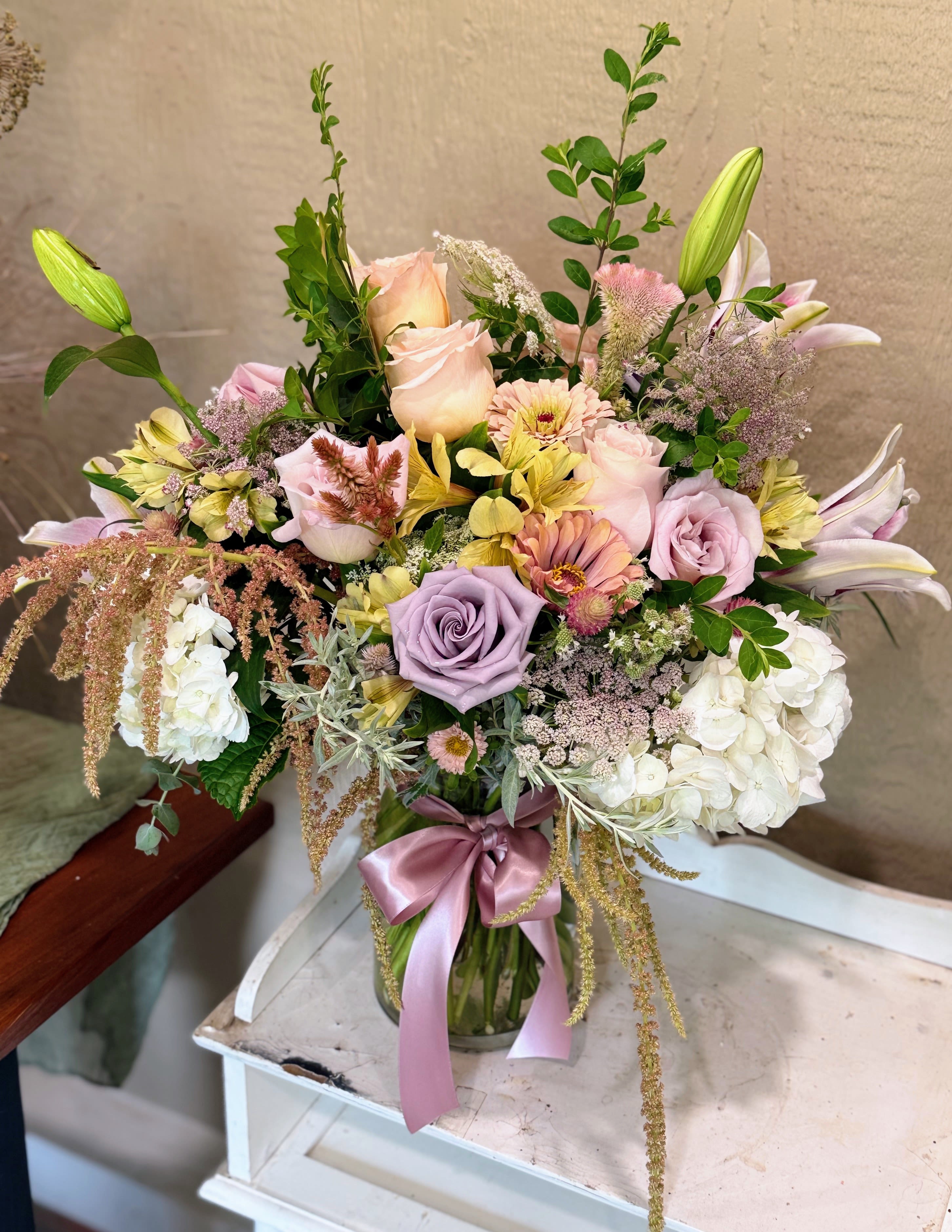 Soft Summertime  - A lush, pastel bouquet that reads like a sunlit garden-this Soft Summer Time flower arrangement is a large mixed bouquet perfect for a birthday, anniversary, spring celebration, or a meaningful everyday gift. The romantic palette of pink, white, lavender, and soft yellow makes the recipient feel gently celebrated and seen, the kind of arrangement that brightens a kitchen table or a bedside with calm, cheerful elegance.  Built in a clear glass vase and finished with a satin pink ribbon, this arrangement features prominent roses and lilies paired with billowy hydrangea, zinnias, draping amaranth, all layered with airy filler and tall greenery for a full, textured silhouette. The composition reads soft and abundant-rosy petals, smooth lily blooms, and cushioned hydrangea create both visual depth and a pleasing contrast of shapes and scents.  Ideal for the person who appreciates classical, feminine florals, Soft Summer Time works beautifully as a thoughtful pick-me-up or a special-occasion centerpiece. The vase makes it ready to display right away; keep blooms in fresh water, away from direct sun and drafts, and trim stems slightly every few days to extend their life.