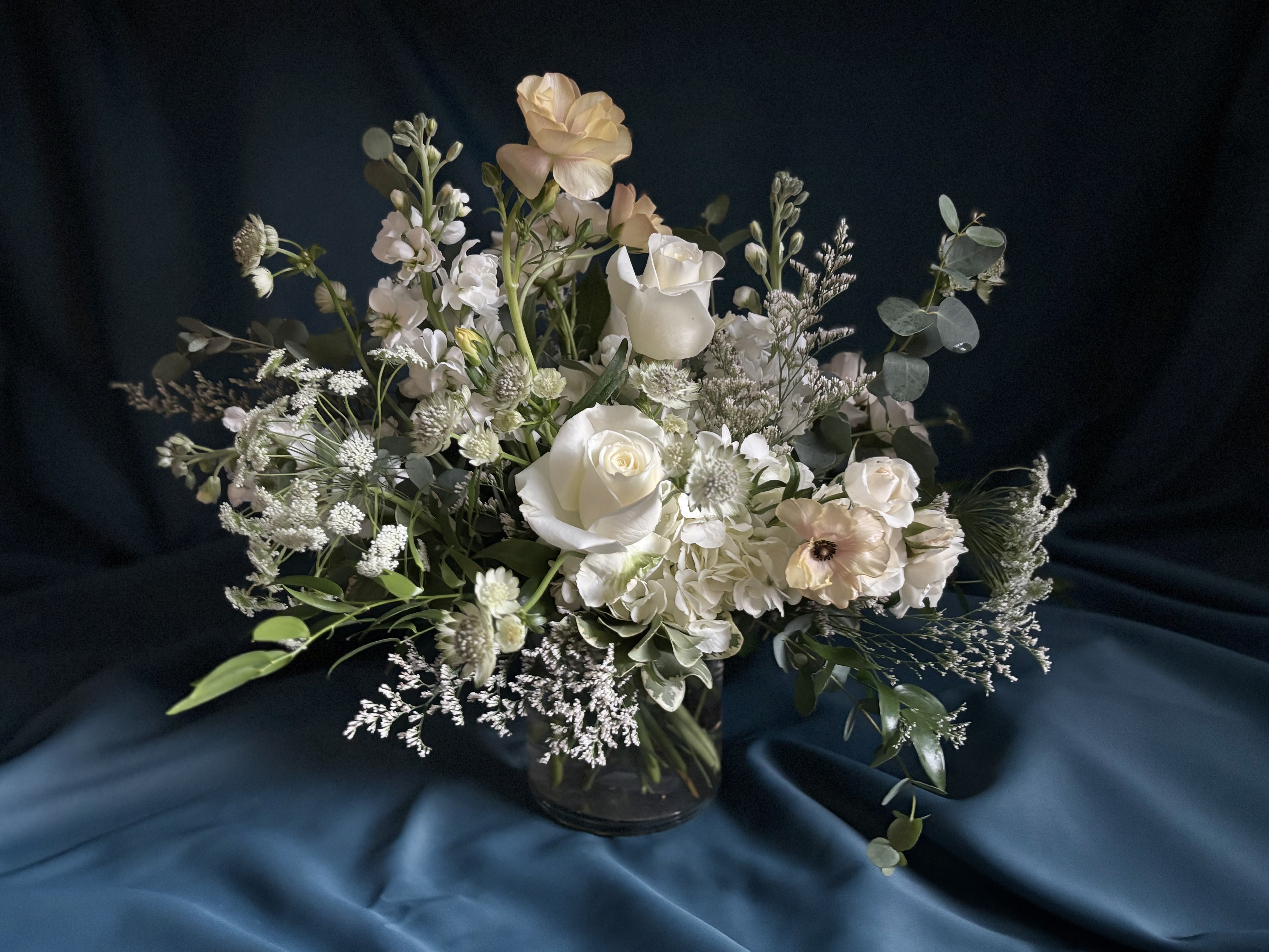 Asymmetrical arrangement of white and cream flowers in a clear glass vase on a dark fabric backdrop