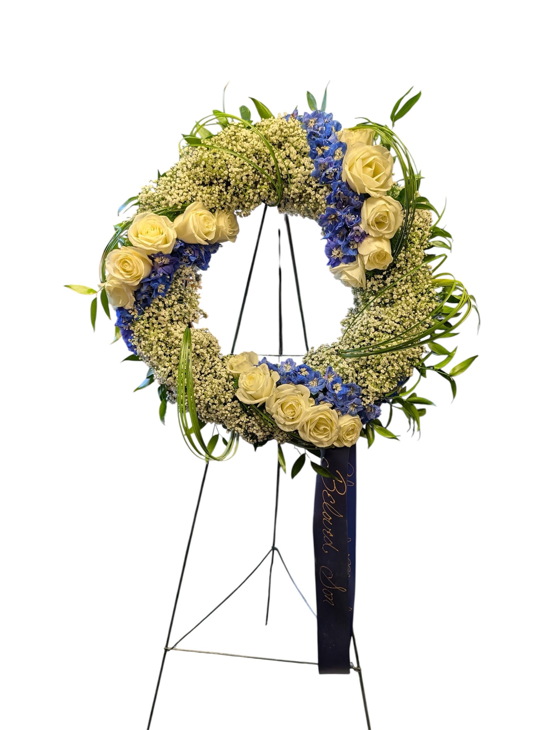 Standing wreath of cream roses, blue blooms, and white filler flowers on an easel with a dark ribbon