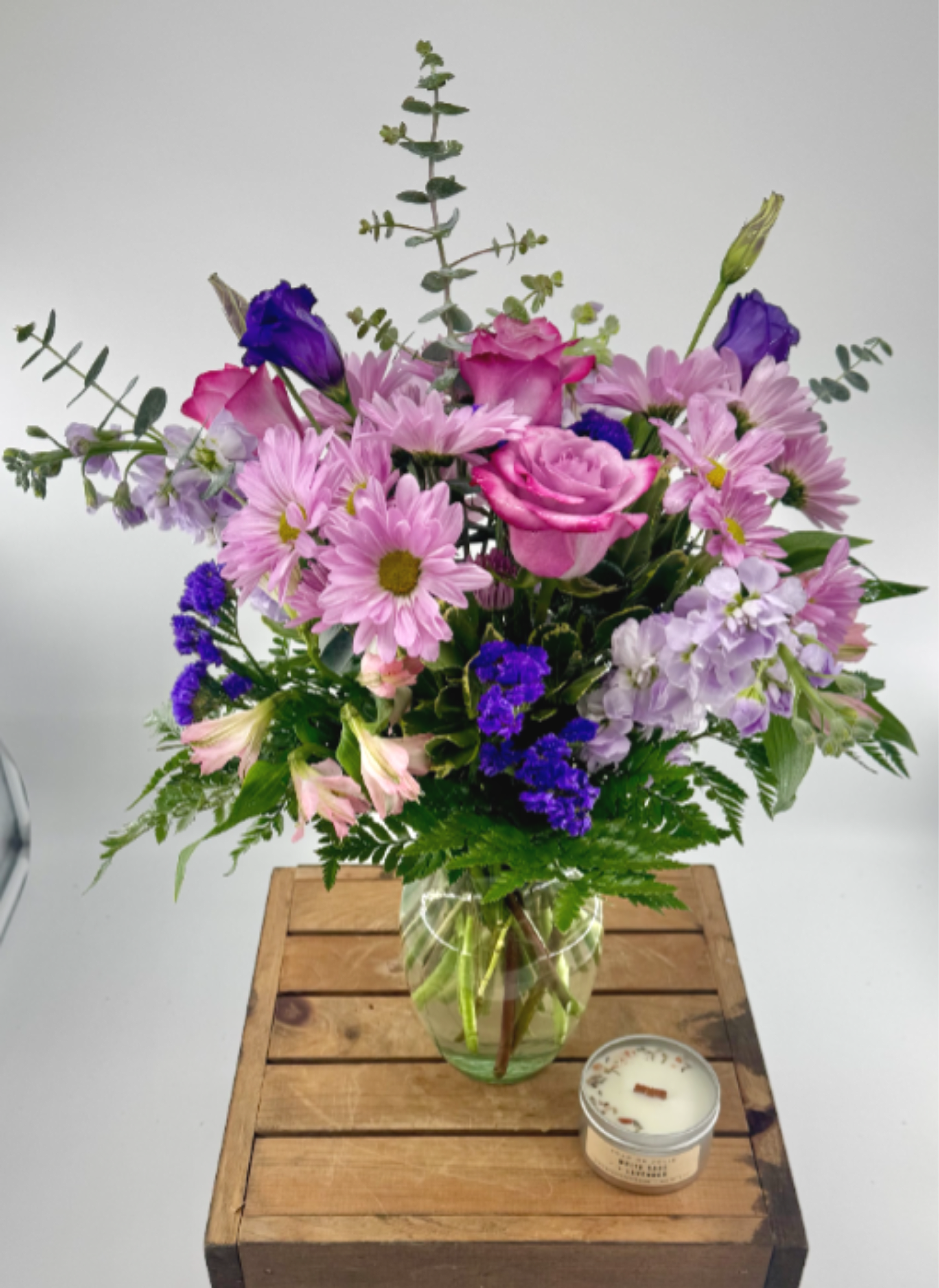 Pink and purple mixed flower bouquet in a clear glass vase on a wood crate with a small tin candle beside it