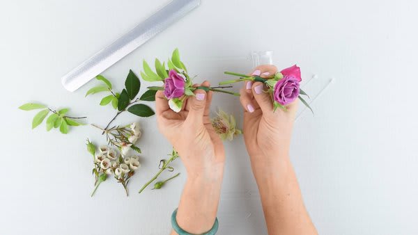 Hands arranging small purple roses with greenery on a white surface