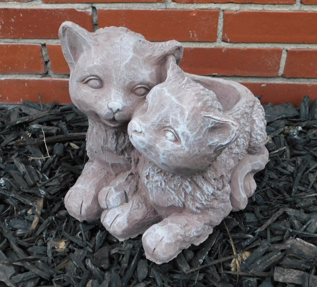 Two small cat figurines sitting together on mulch in front of a brick wall.