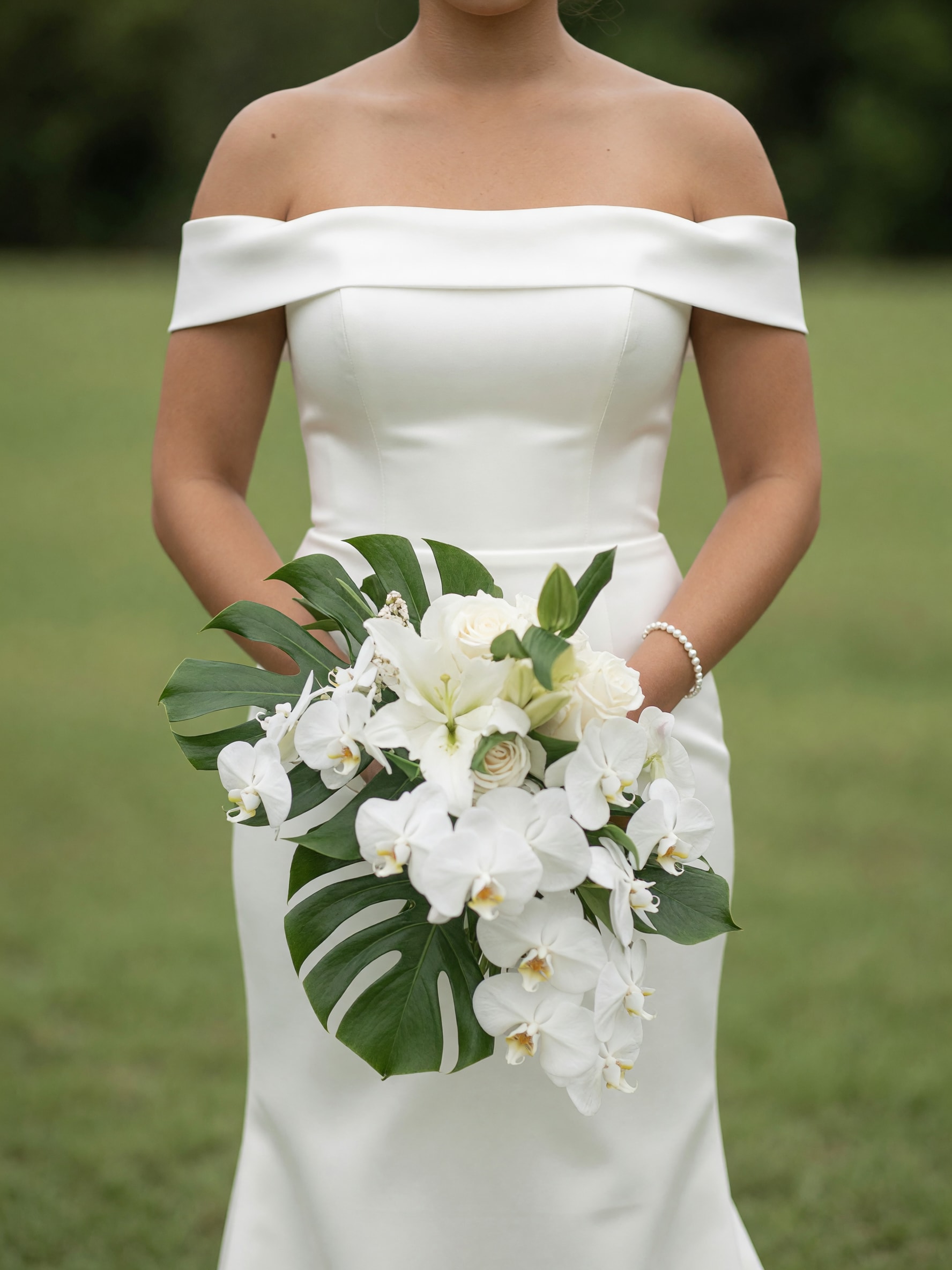 Island Elegance™ Cascading Orchid Bridal Bouquet - Walk down the aisle carrying timeless sophistication with our Island Elegance™ Cascading Orchid Bridal Bouquet. This breathtaking design features cascading white Phalaenopsis orchids, fragrant white lilies, premium white roses, and lush tropical monstera foliage, creating a graceful bouquet that is both modern and unforgettable.  Inspired by tropical luxury and contemporary bridal style, this bouquet is perfect for destination weddings, elegant ballroom celebrations, garden ceremonies, and beachfront vows. The flowing orchids symbolize beauty, purity, and everlasting love, while the clean white palette creates a classic look that never goes out of style.  Each bouquet is handcrafted by the award-winning designers at Tommy Austin Florist, using premium flowers selected for their beauty, freshness, and elegance  