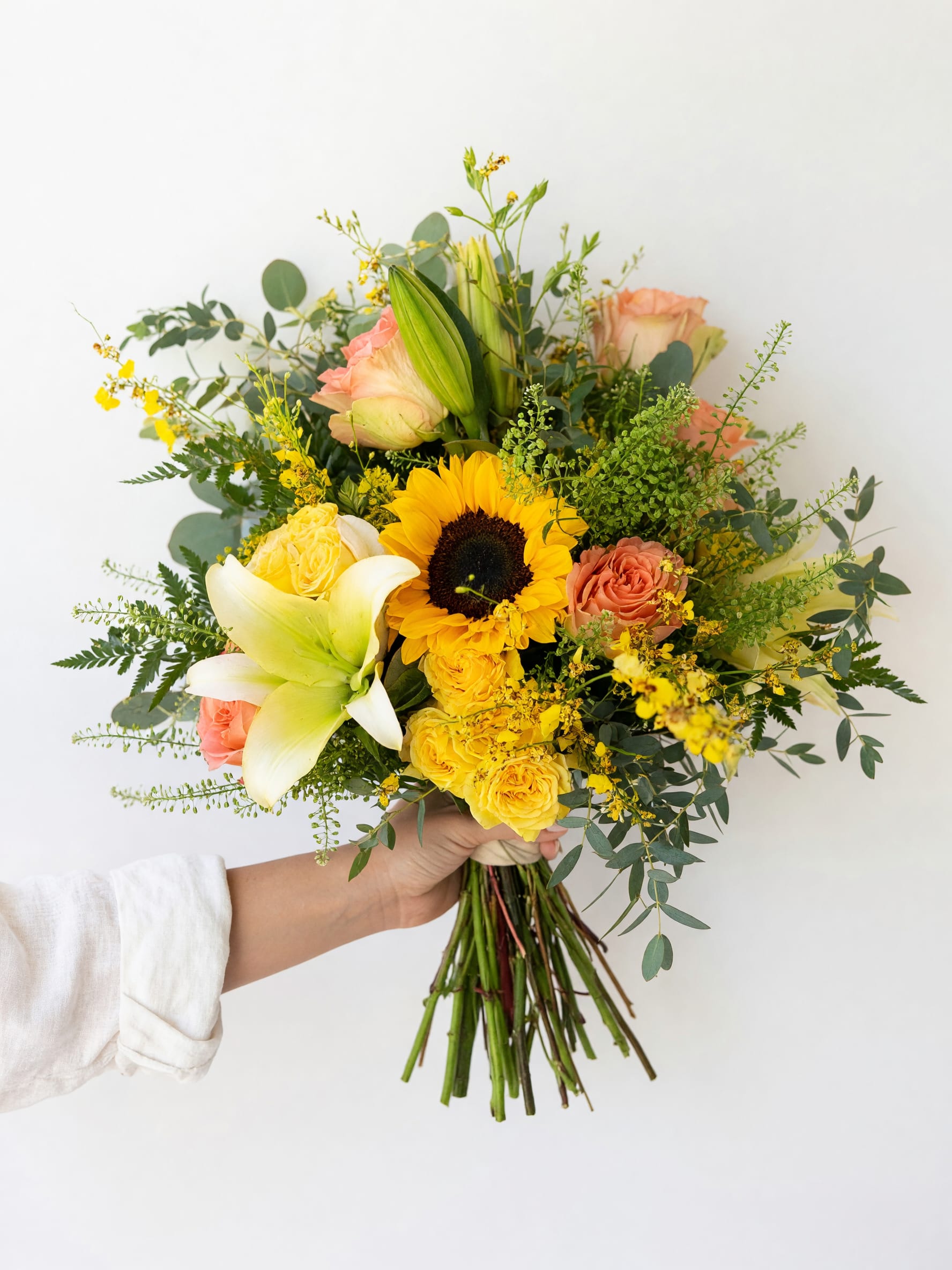 Golden Garden™ Hand-Tied Bouquet - Bring sunshine and elegance together with our Golden Garden™ Hand-Tied Bouquet. Designed with a vibrant sunflower, fragrant white lily, soft peach roses, cheerful yellow spray roses, delicate dancing orchids, fresh eucalyptus, and lush seasonal greenery, this bouquet is hand-tied in a relaxed garden style that’s full of texture and natural beauty.  Perfect for birthdays, anniversaries, congratulations, thank-you gifts, get well wishes, or simply brightening someone’s day, this bouquet is handcrafted with premium fresh flowers and wrapped for a beautiful presentation.  Every bouquet is expertly designed by Tommy Austin Florist using the freshest blooms available to ensure a unique and memorable gift. Each bouquet is handcrafted using fresh seasonal flowers. Flower varieties may vary slightly depending on availability while maintaining the same beautiful color palette, style, and value.