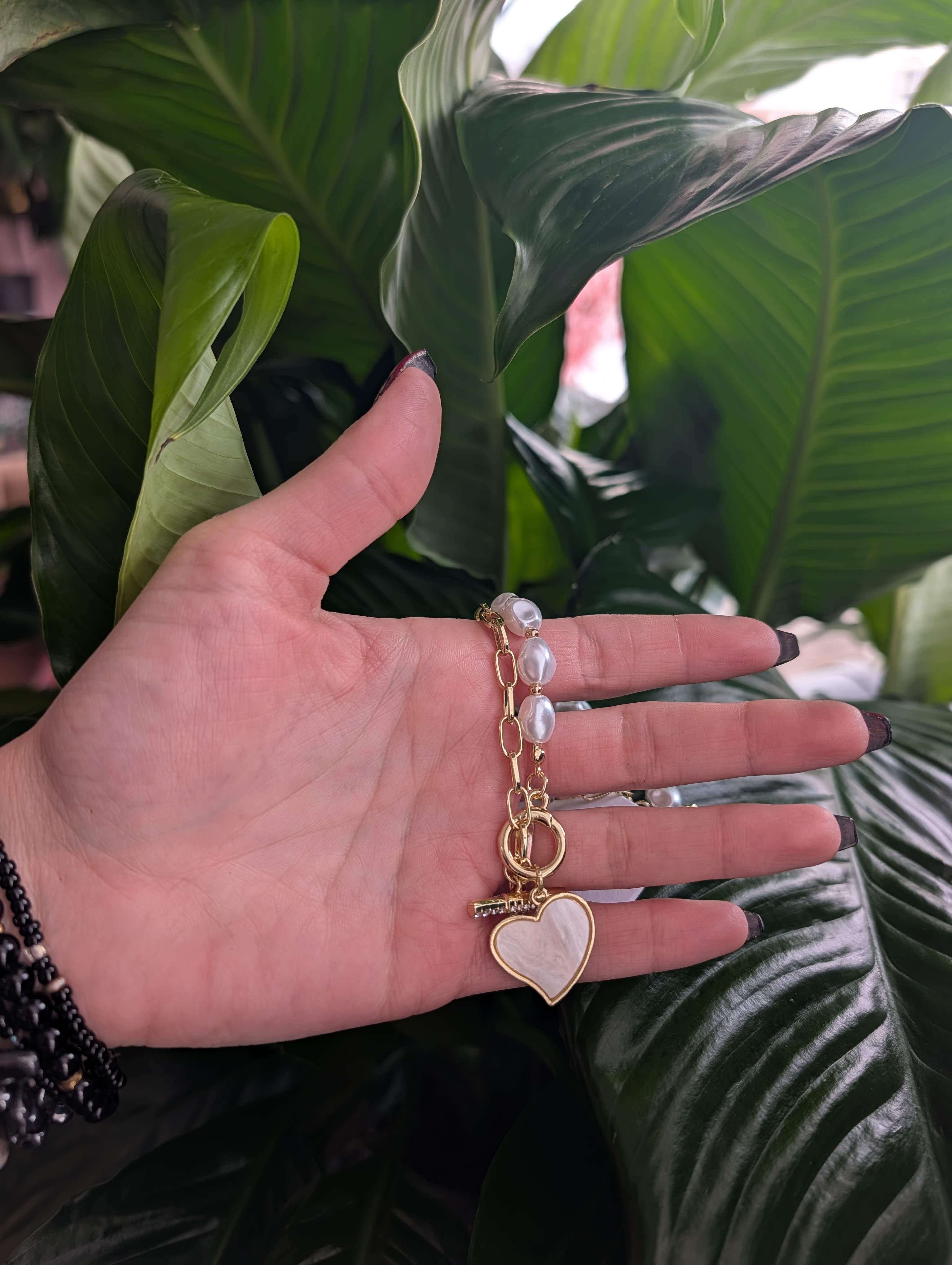 Hand holding a gold heart keychain in front of large green leaves