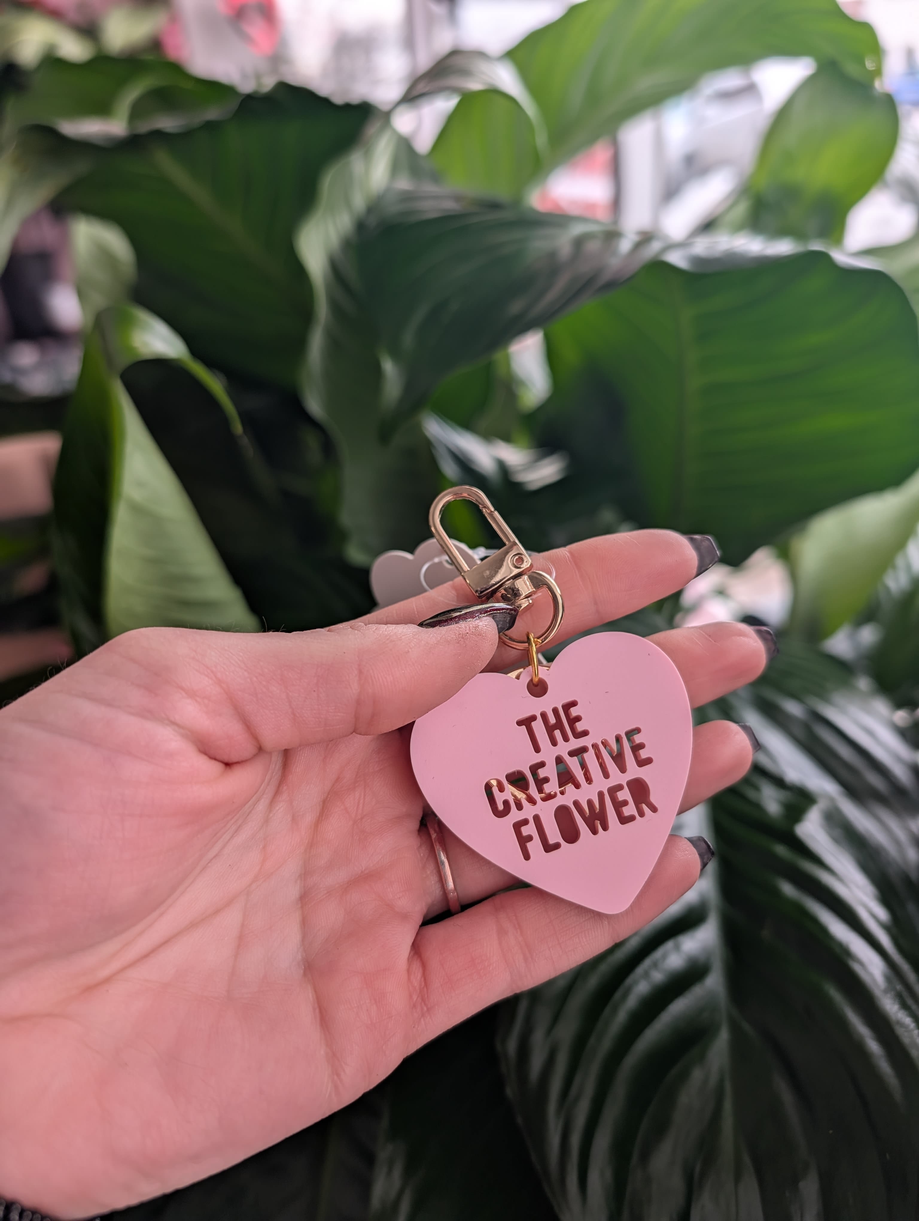Hand holding a pink heart keychain in front of large green leaves