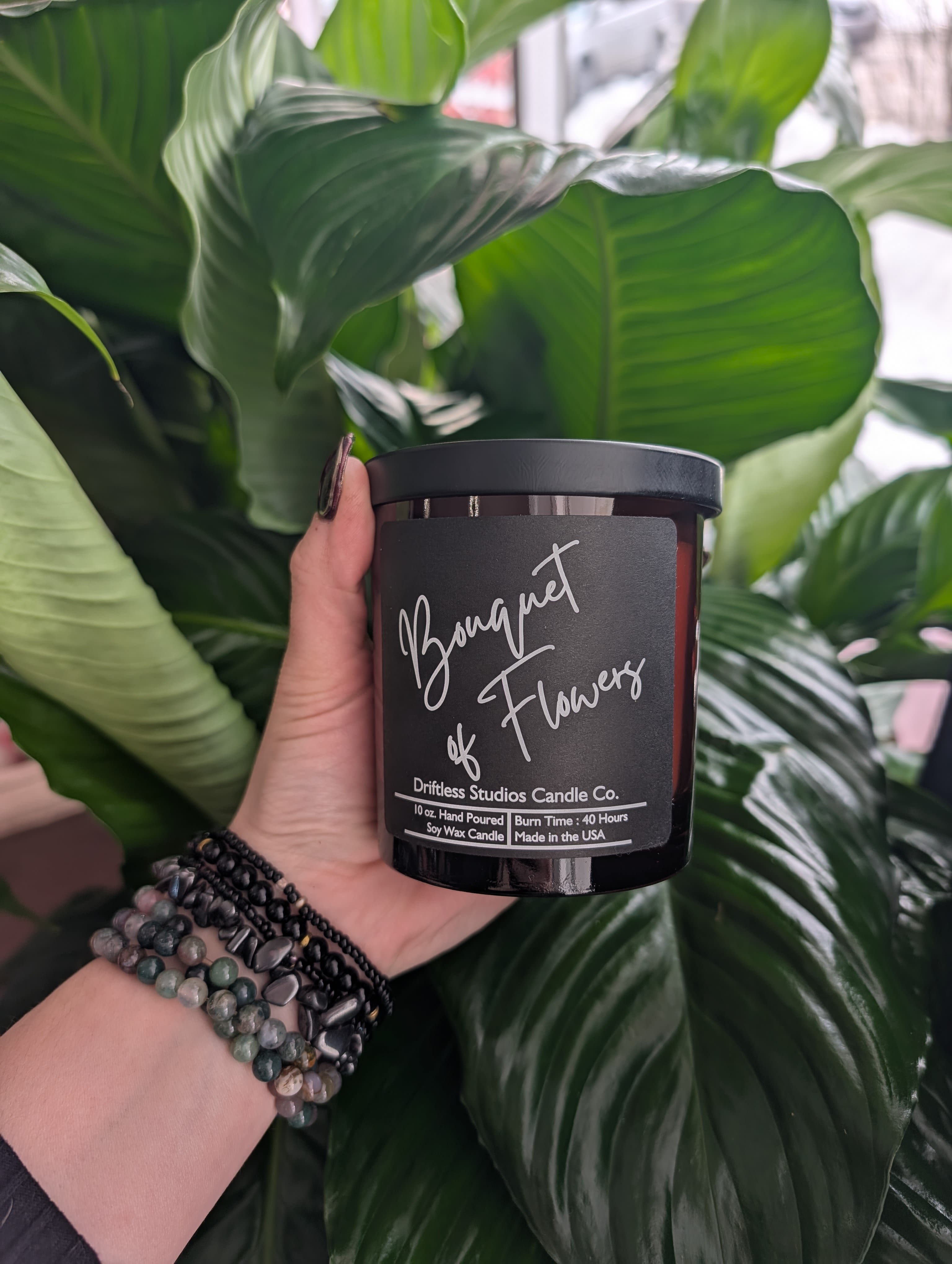 Hand holding a black candle jar labeled "Bouquet of Flowers" in front of large green leaves.