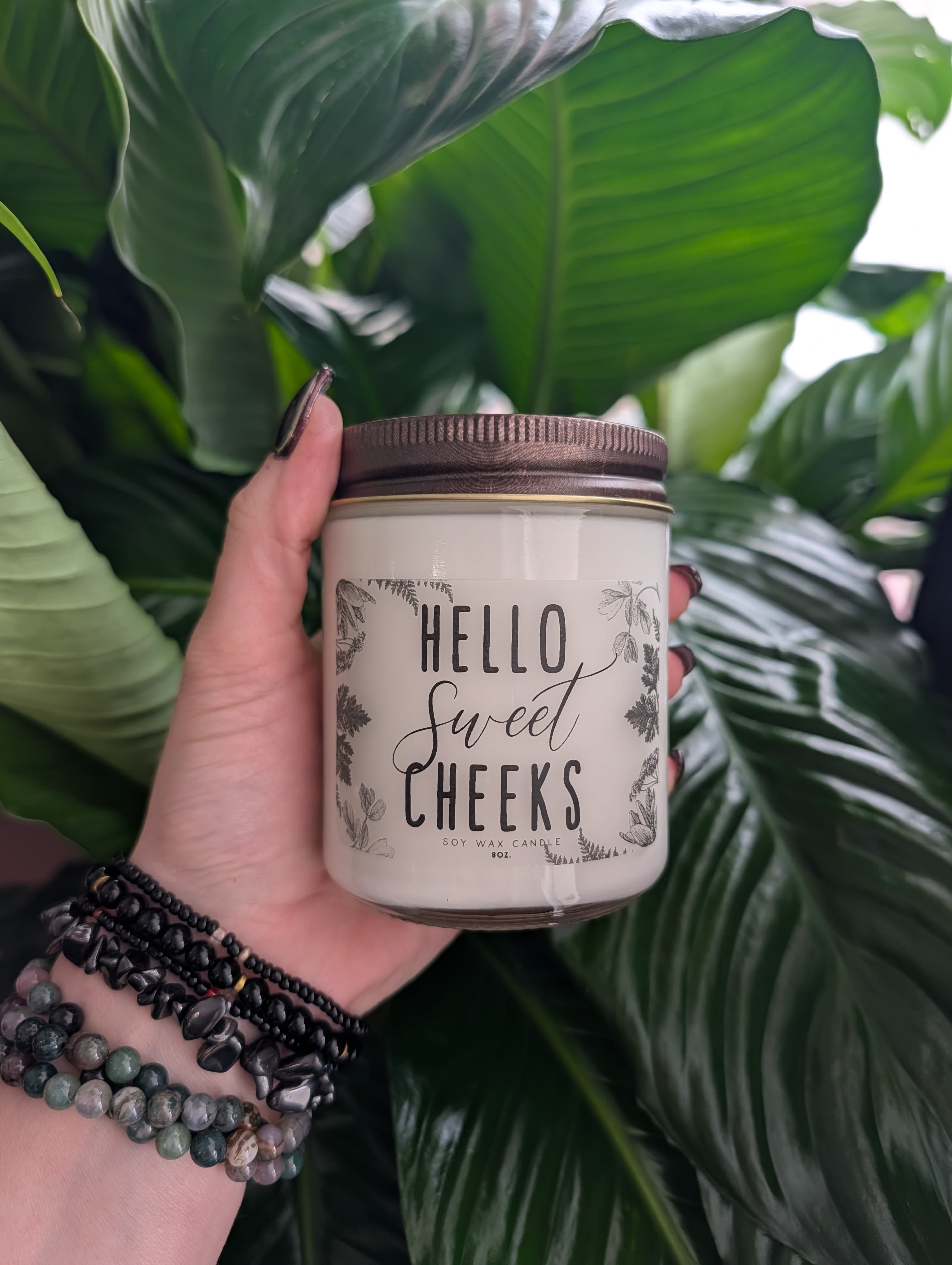 Hand holding a soy wax candle jar in front of large green leaves