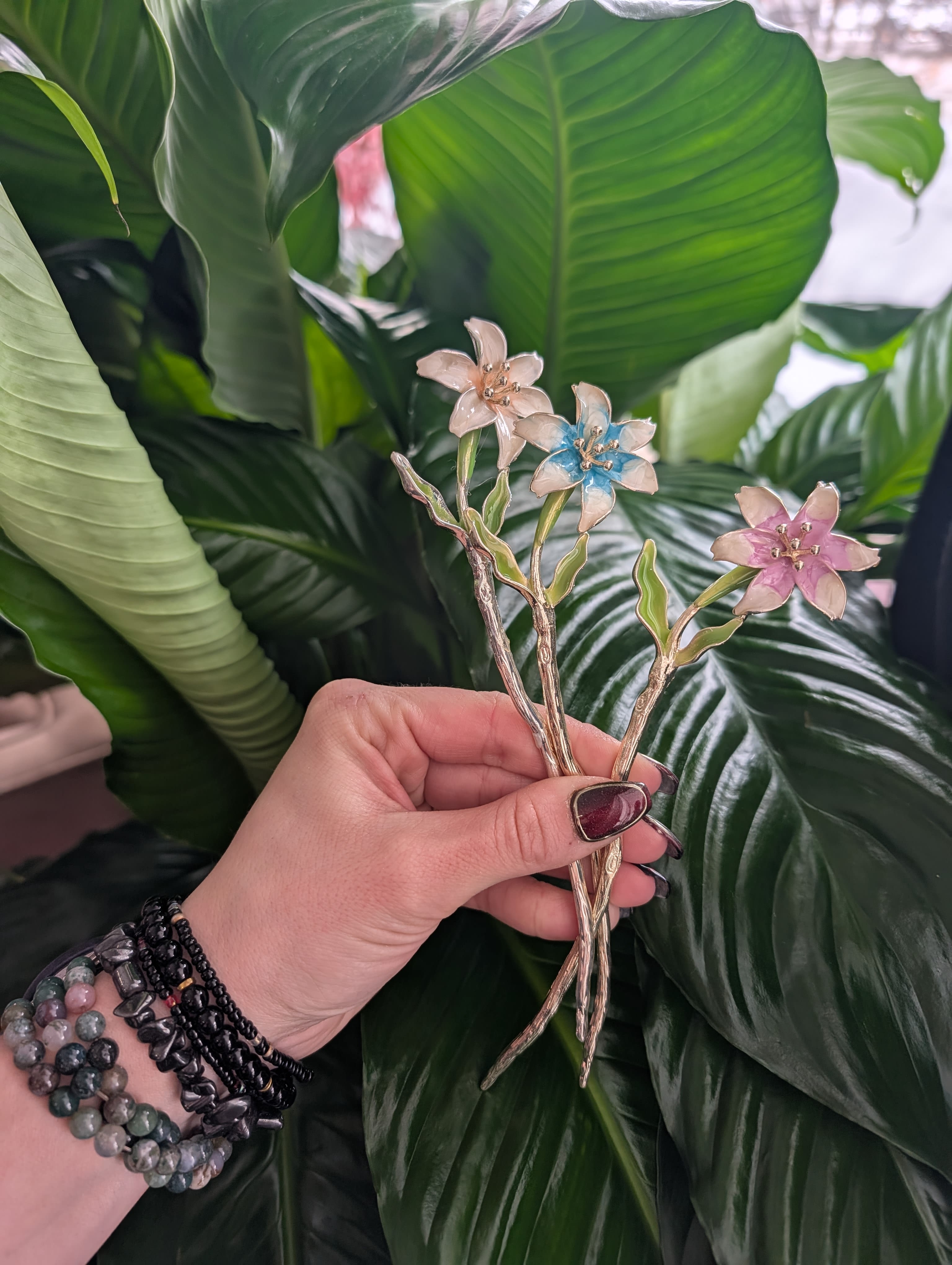 Hand holding three small enamel flower pins against large green leaves