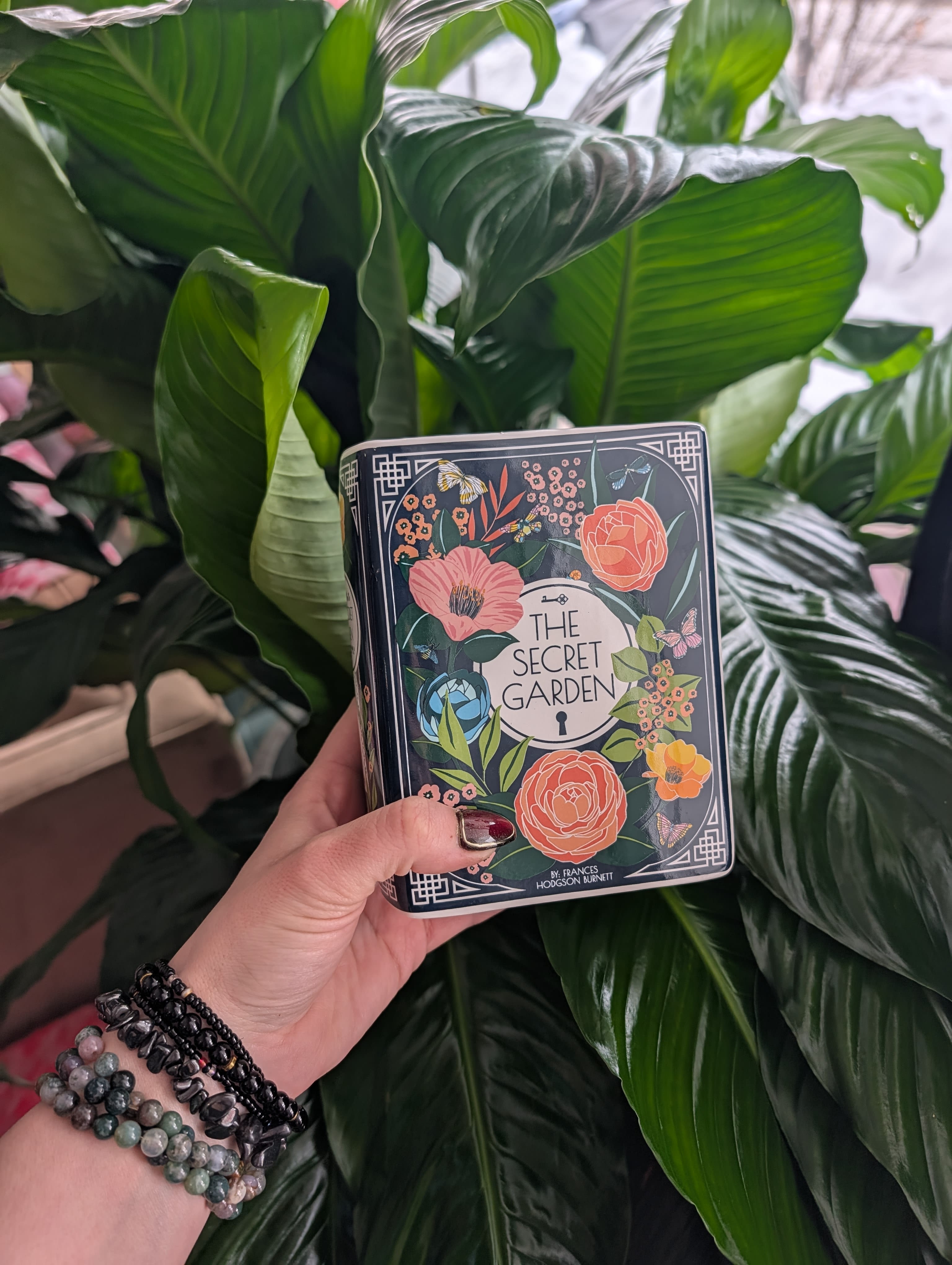 Hand holding a decorative tin box in front of large green leaves