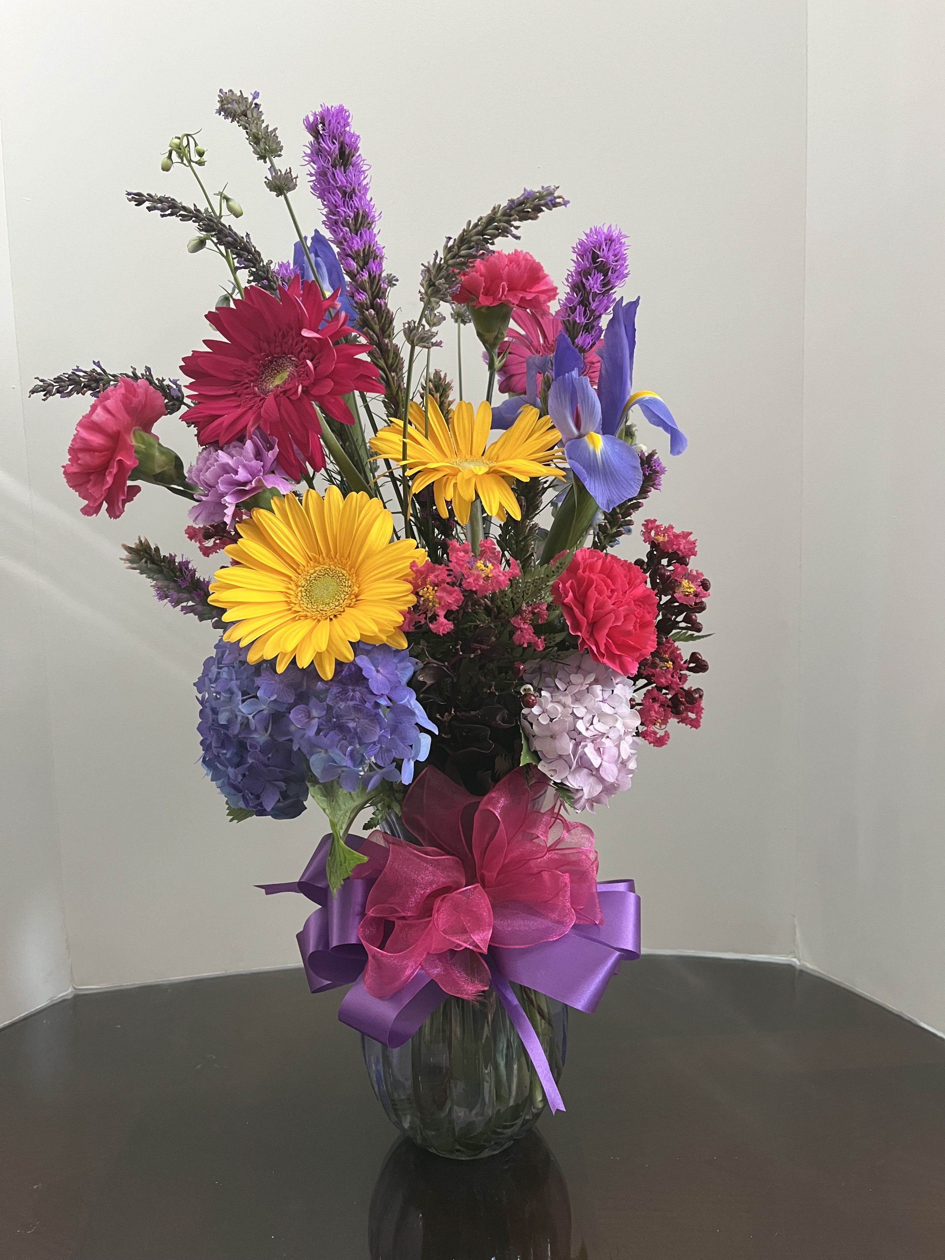 Tall mixed arrangement with yellow gerbera, blue hydrangeas, pink carnations and purple blooms in a glass vase.