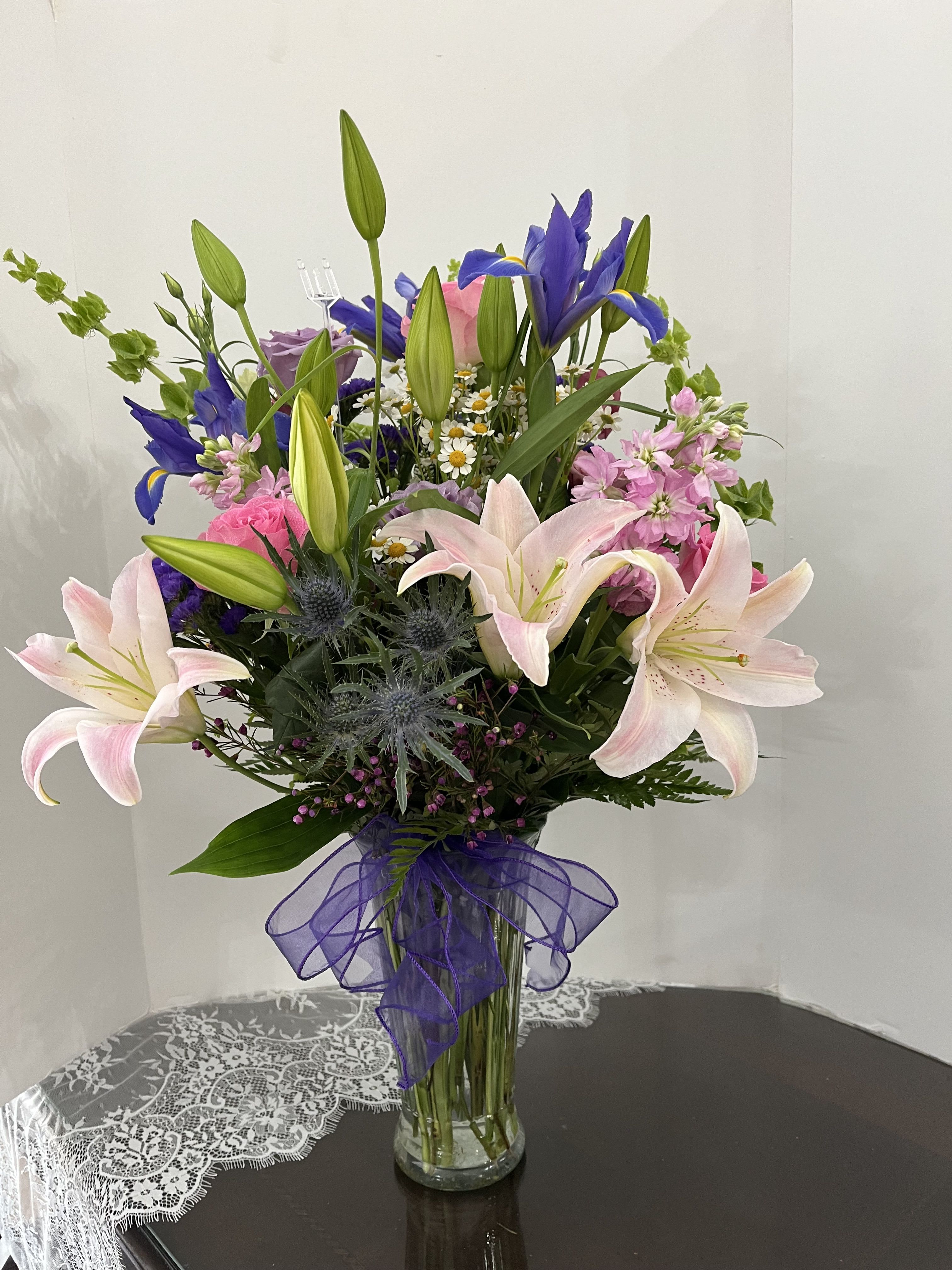 Mixed bouquet of lilies, irises, and pink flowers in a glass vase with a purple ribbon