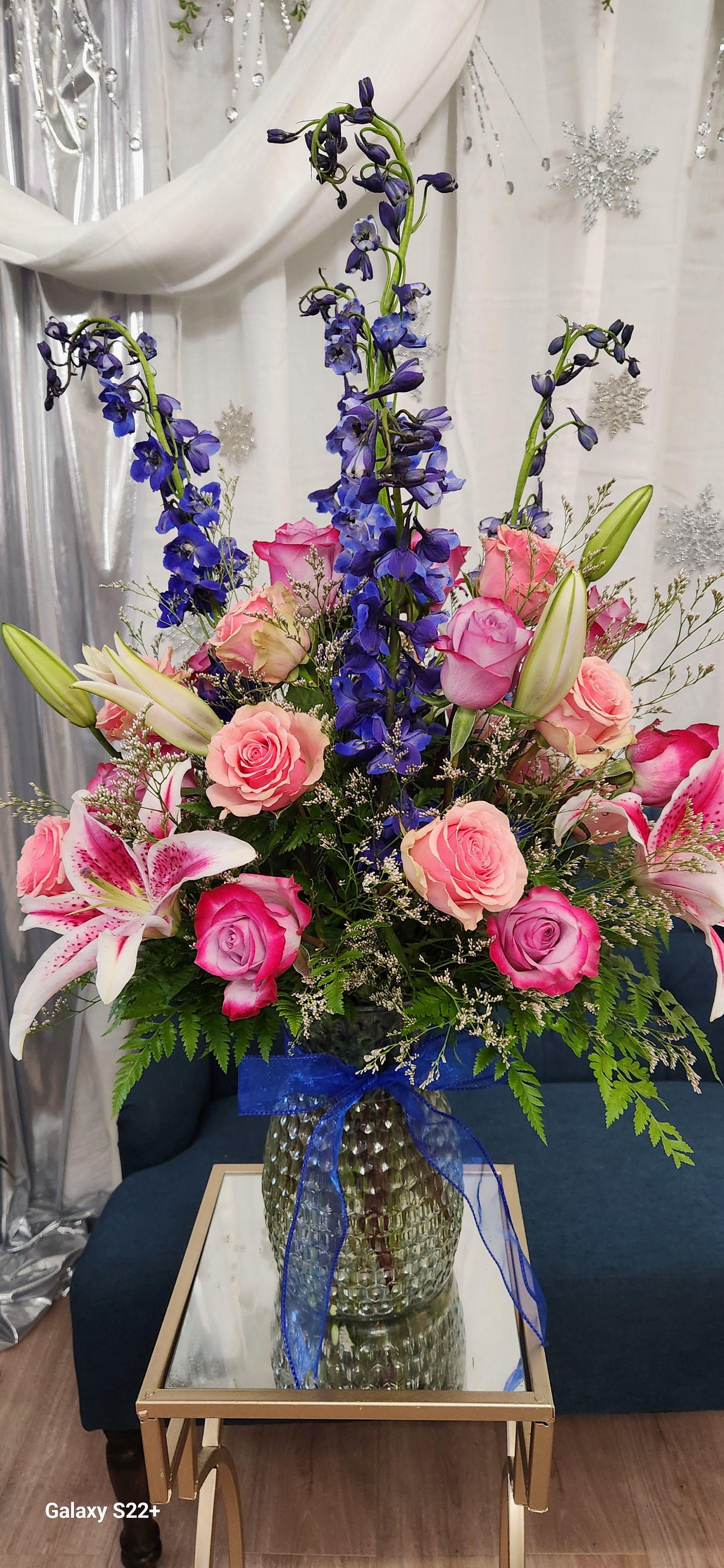 Tall bouquet of pink roses, lilies, and blue delphinium in a glass vase