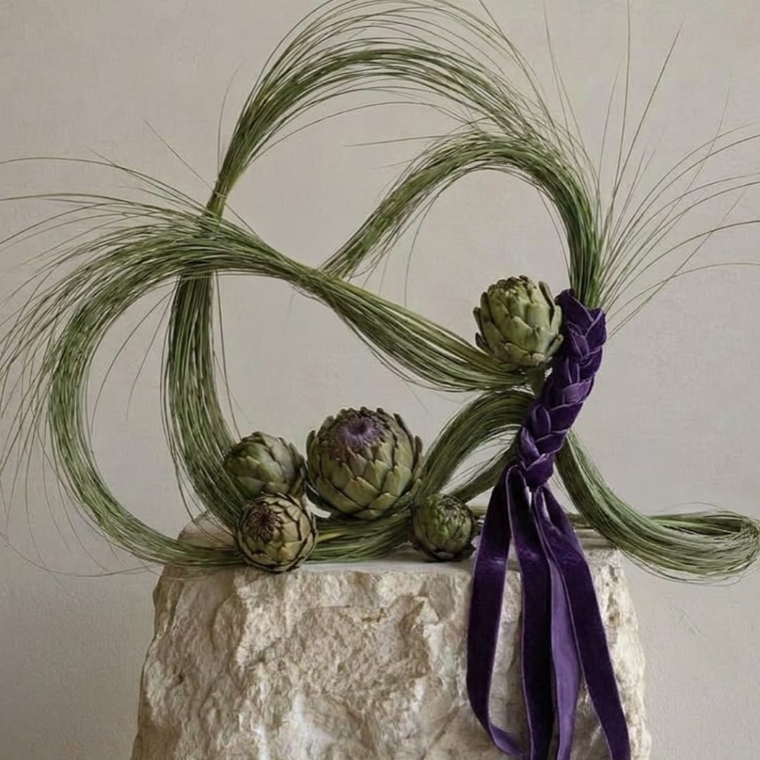 Artistic floral arrangement with green artichokes and looping grass in a textured vase