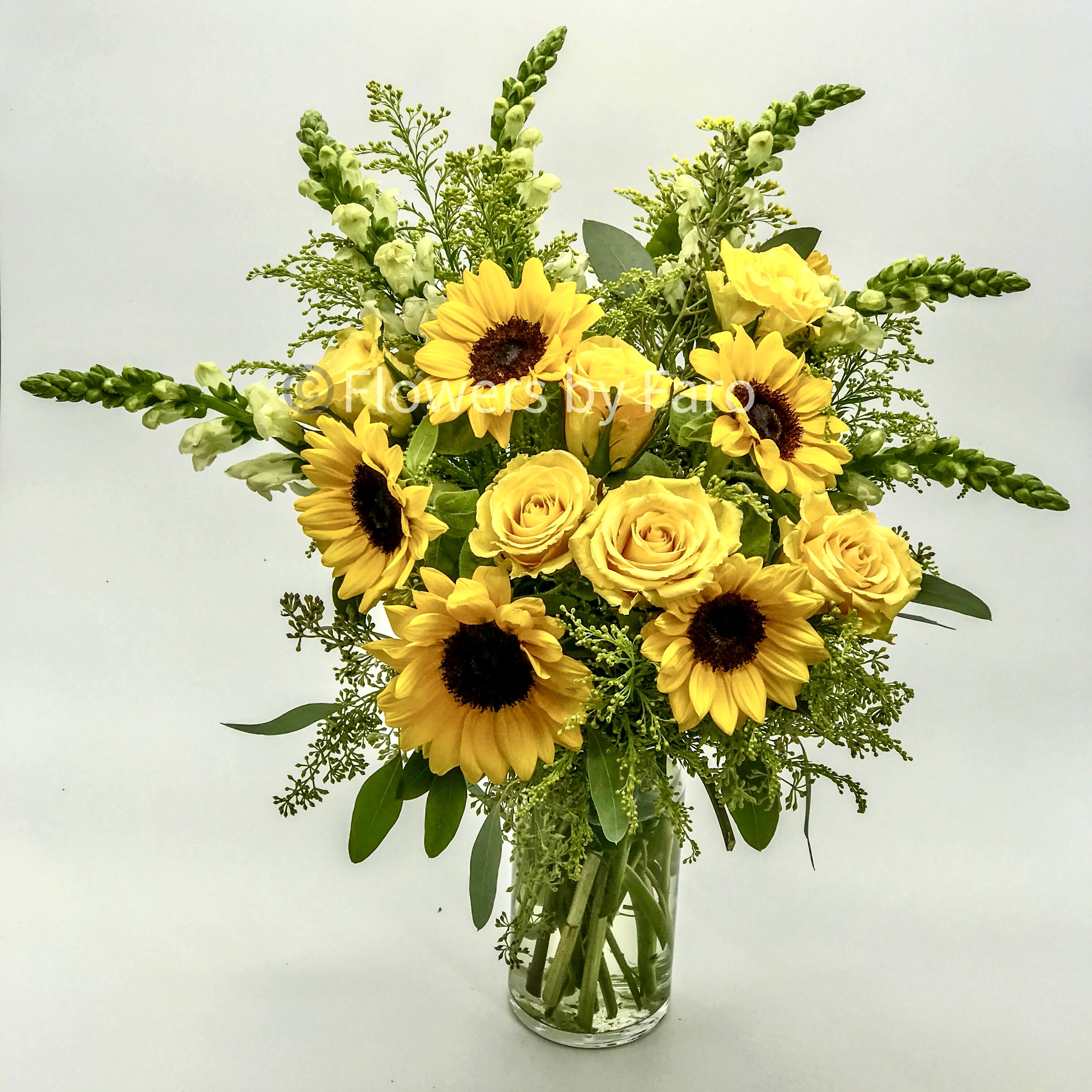 Tall arrangement of yellow sunflowers, yellow roses, and white snapdragons in a clear glass vase