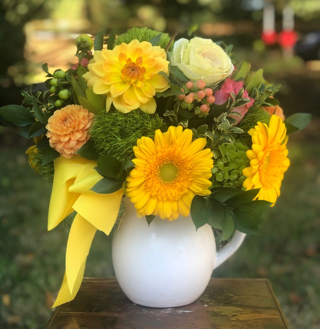 Yellow and peach flowers arranged in a white pitcher vase with a yellow ribbon