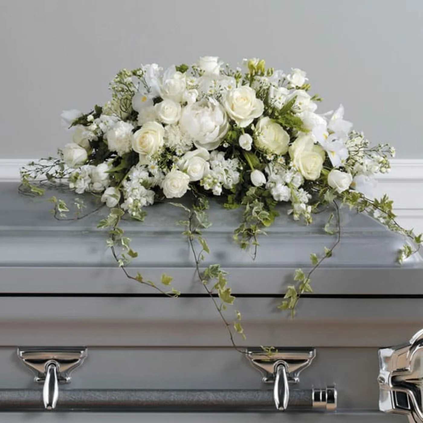 Low white rose and lily arrangement with trailing vines displayed on a silver casket
