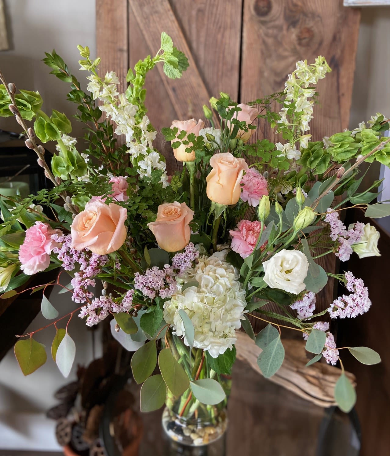 Tall arrangement of peach roses, white hydrangea, pink flowers and green bells of Ireland in a glass vase