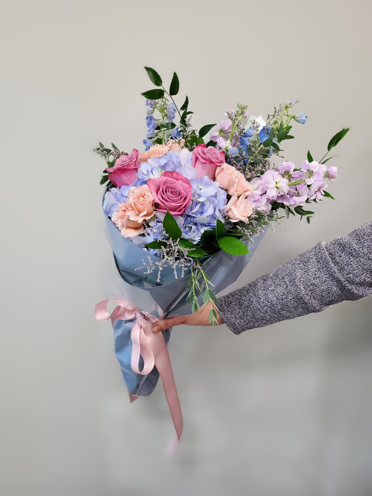 Handheld bouquet of pink roses and pastel blue flowers wrapped in blue paper