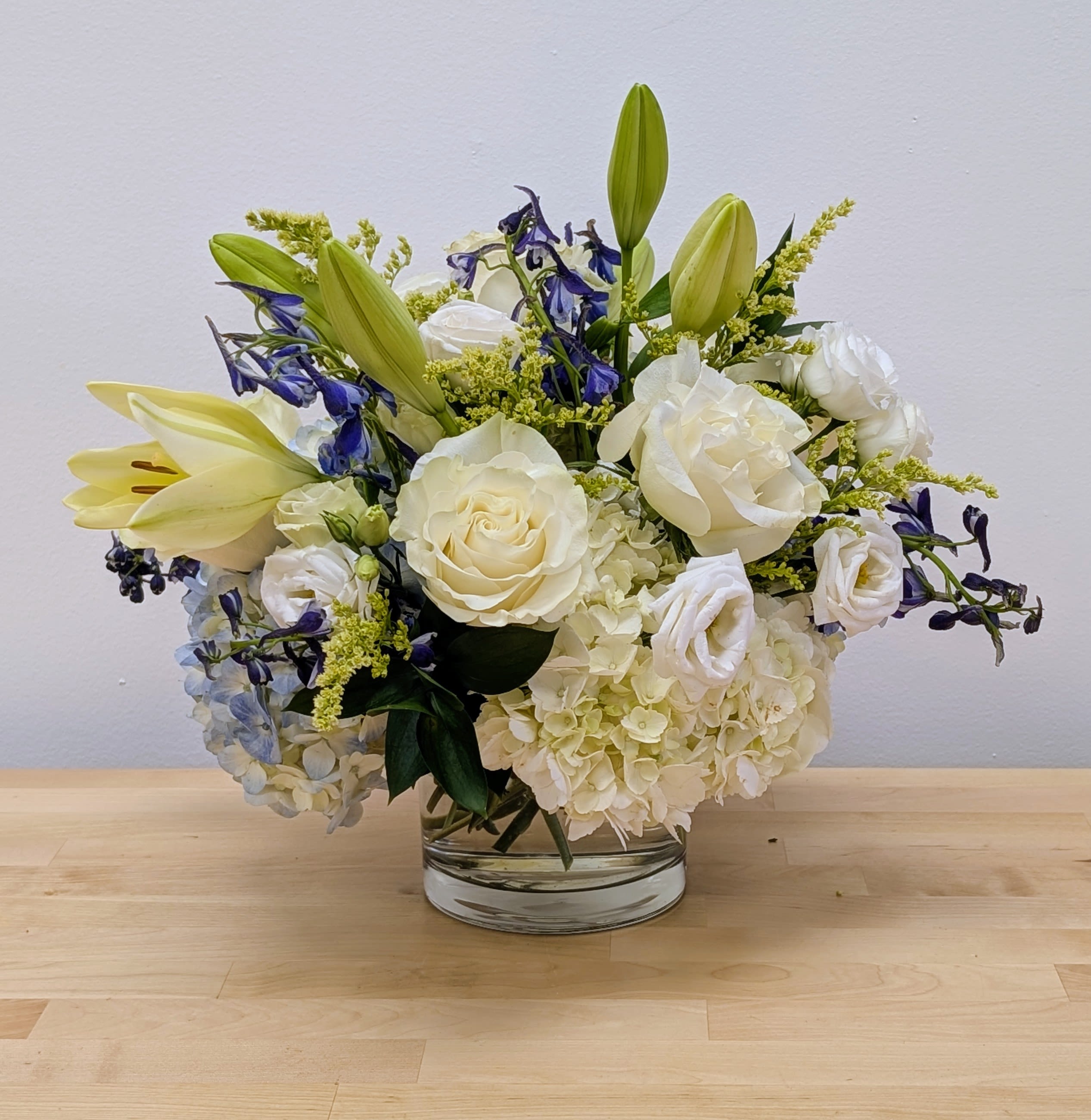 White roses, lilies, and hydrangeas with blue accents arranged in a low clear glass vase