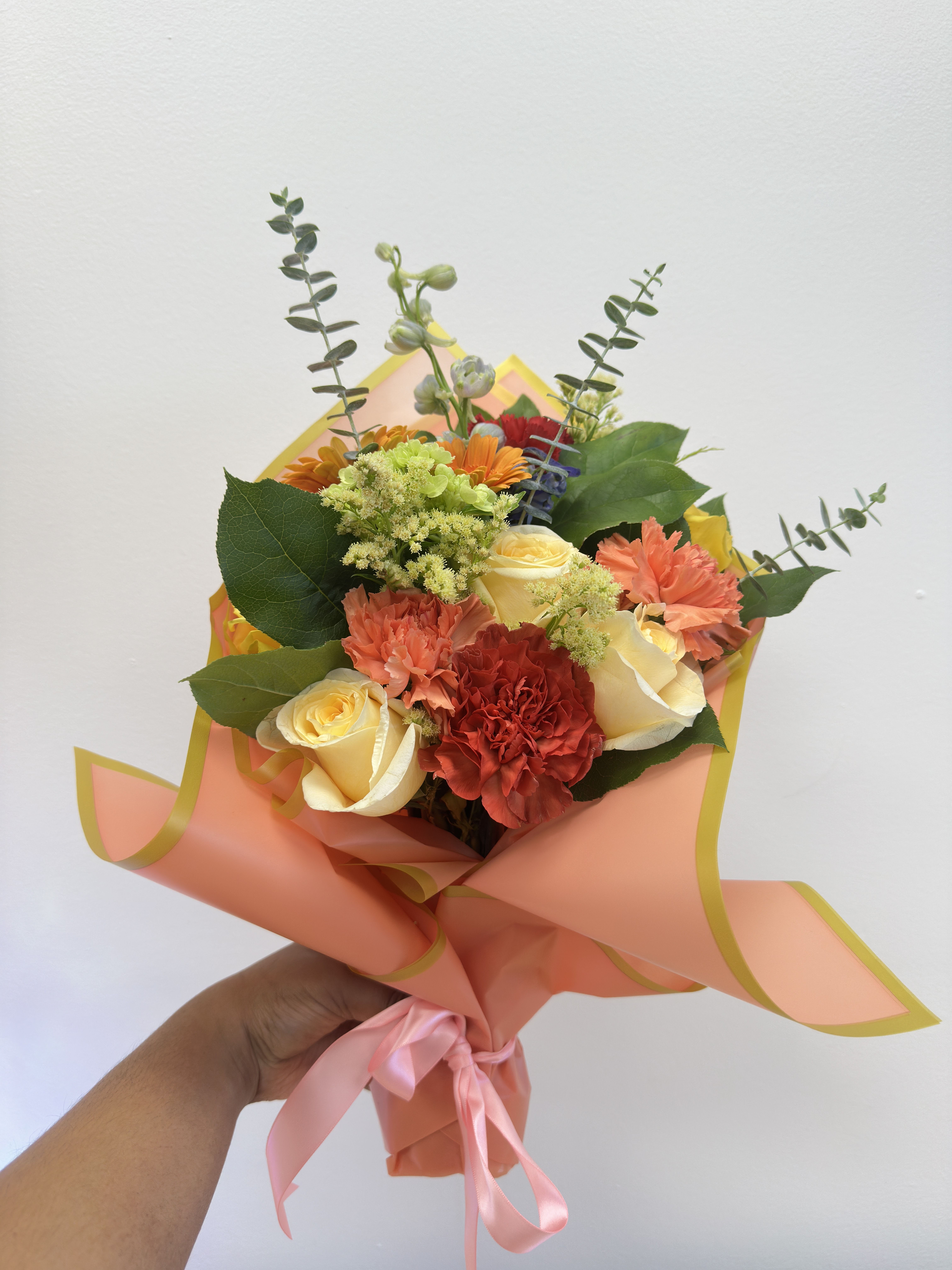 Handheld bouquet of roses, carnations, and orange flowers wrapped in peach paper
