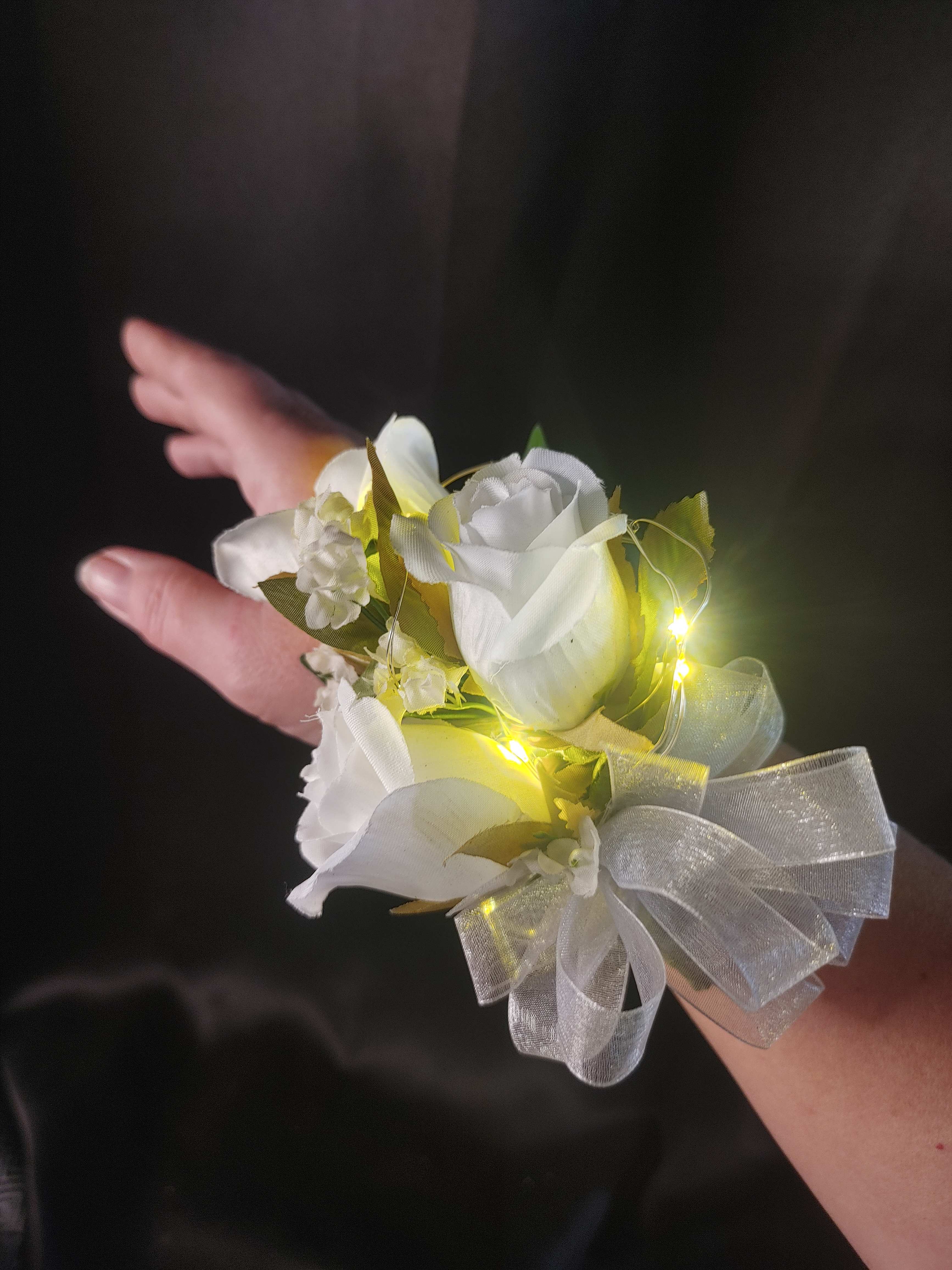 White rose wrist corsage with sheer silver bow and tiny lights on a person’s arm.